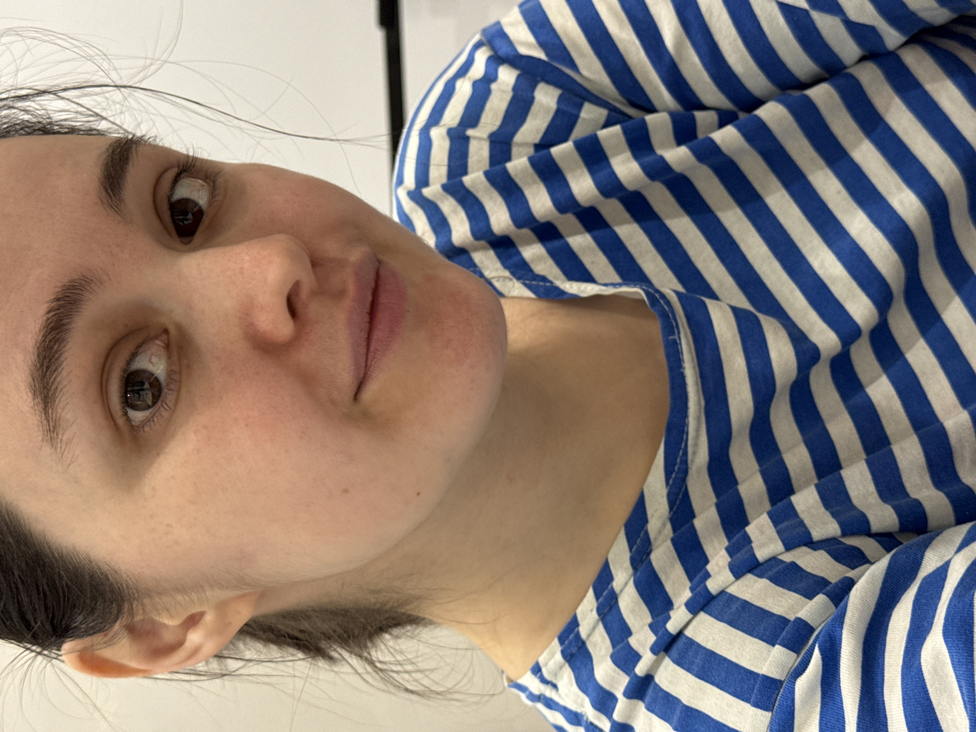 A close-up, canted view depicts a woman wearing a blue and white striped shirt. The focus is on her face, revealing brown eyes, dark eyebrows, and fair skin with some minor blemishes. Some strands of dark hair are visible. The background is a plain white wall. There is no visual information to indicate a specific location, let alone Izhevsk, Russia.