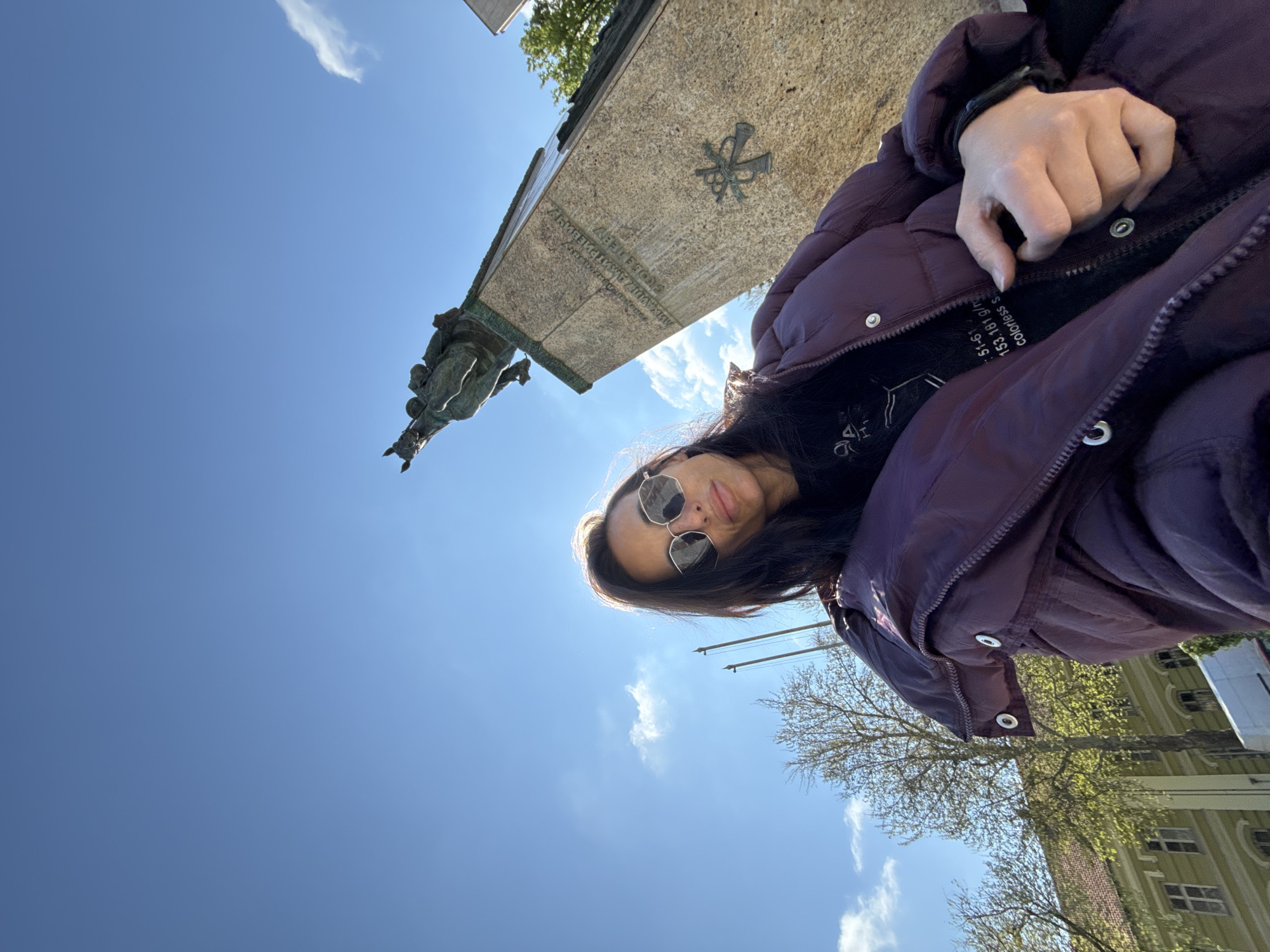 A medium shot captures a woman in a purple down jacket, angled to the right with a bright blue sky behind her. She's wearing sunglasses and is positioned near a monument. The monument includes a bronze equestrian statue on top of a stone base. The base bears a cross-like symbol. Trees and a light yellow building are visible on the right side of the frame. The setting is Pleven, Bulgaria.