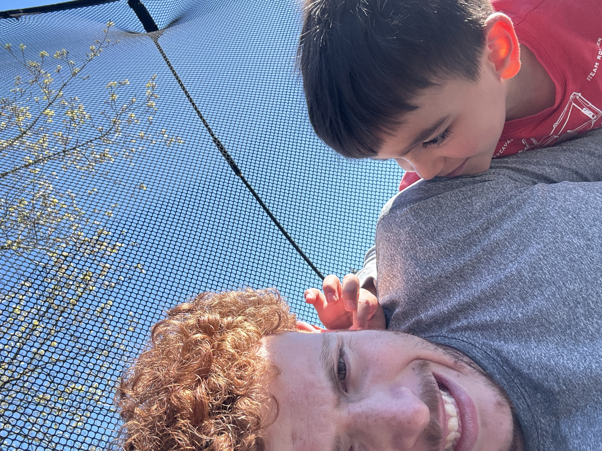 The image shows a man with red curly hair lying on his back on a trampoline. A young boy with dark hair is lying on top of the man, his face close to the man's neck. The trampoline netting is visible in the background, along with a blue sky and tree branches with budding leaves. The location is likely a residential area, and it is possible that the image was taken in the city of Pepper Pike, United States of America.