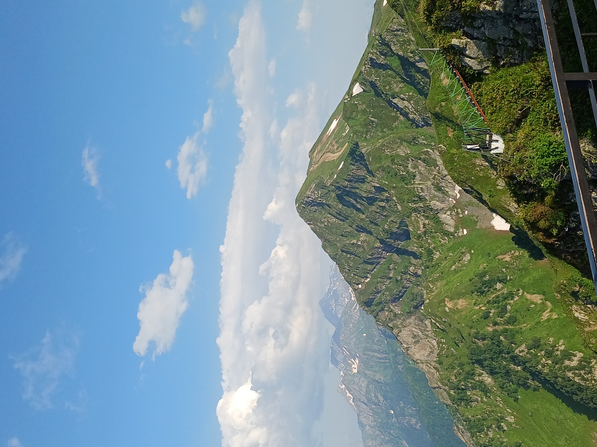 The image depicts a scenic mountain landscape with lush green slopes under a bright blue sky dotted with white clouds. In the foreground, a dark metal railing runs horizontally across the bottom of the frame. Behind the railing, a steep, vegetated mountainside is visible. Further in the background, a prominent, jagged green mountain with patches of snow dominates the center of the image. To the left and behind this central peak, a range of more distant, rugged mountains with snow-capped summits can be seen. A small, white structure with a green, zig-zagging bridge or walkway is situated on a rocky outcrop on the right side of the central mountain. The scene is likely captured from a high vantage point, offering a panoramic view of the mountainous terrain. This type of landscape is characteristic of resort areas like Estosadok, Russia, which is known for its mountain ranges and ski infrastructure.