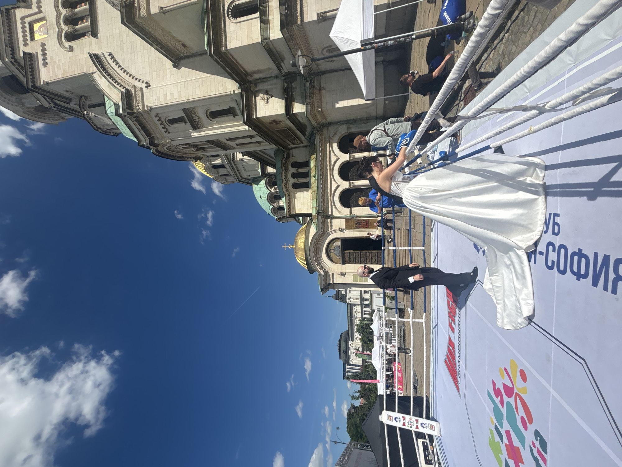 A wedding ceremony unfolds in a boxing ring, set against the backdrop of the Alexander Nevsky Cathedral in Sofia, Bulgaria. The bride, in a white gown, stands inside the ring near the ropes. Her groom, in a black suit, stands alongside her. The ring is set on a white canvas printed with colorful graphics and text, and the cathedral's domes and facade loom in the background. A bright blue sky is visible above, partially filled with scattered clouds. Other people are seen outside the ring, seemingly part of the event or observing it.