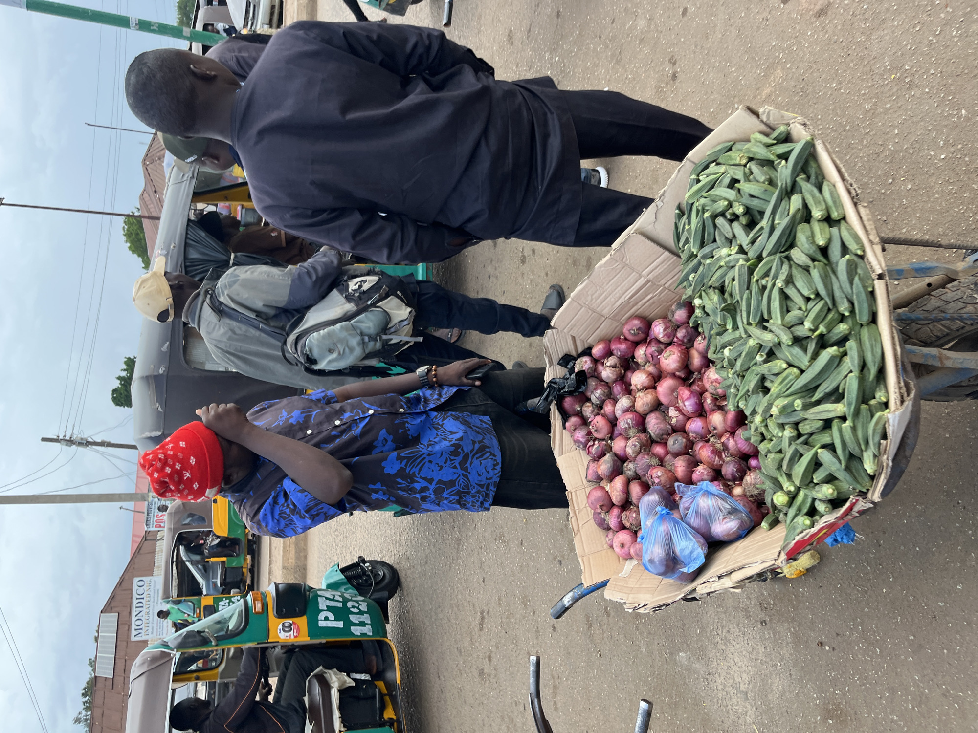 In Bukuru, Nigeria, a man wearing a red beanie and a blue patterned shirt stands with his hand to his forehead, appearing to be in distress. In front of him, a cart is filled with a large quantity of fresh red onions and okra. Other individuals are present in the background, some near a green and yellow auto-rickshaw. The scene appears to be a street market or transportation hub, with paved ground and buildings visible in the distance. The sky is overcast.