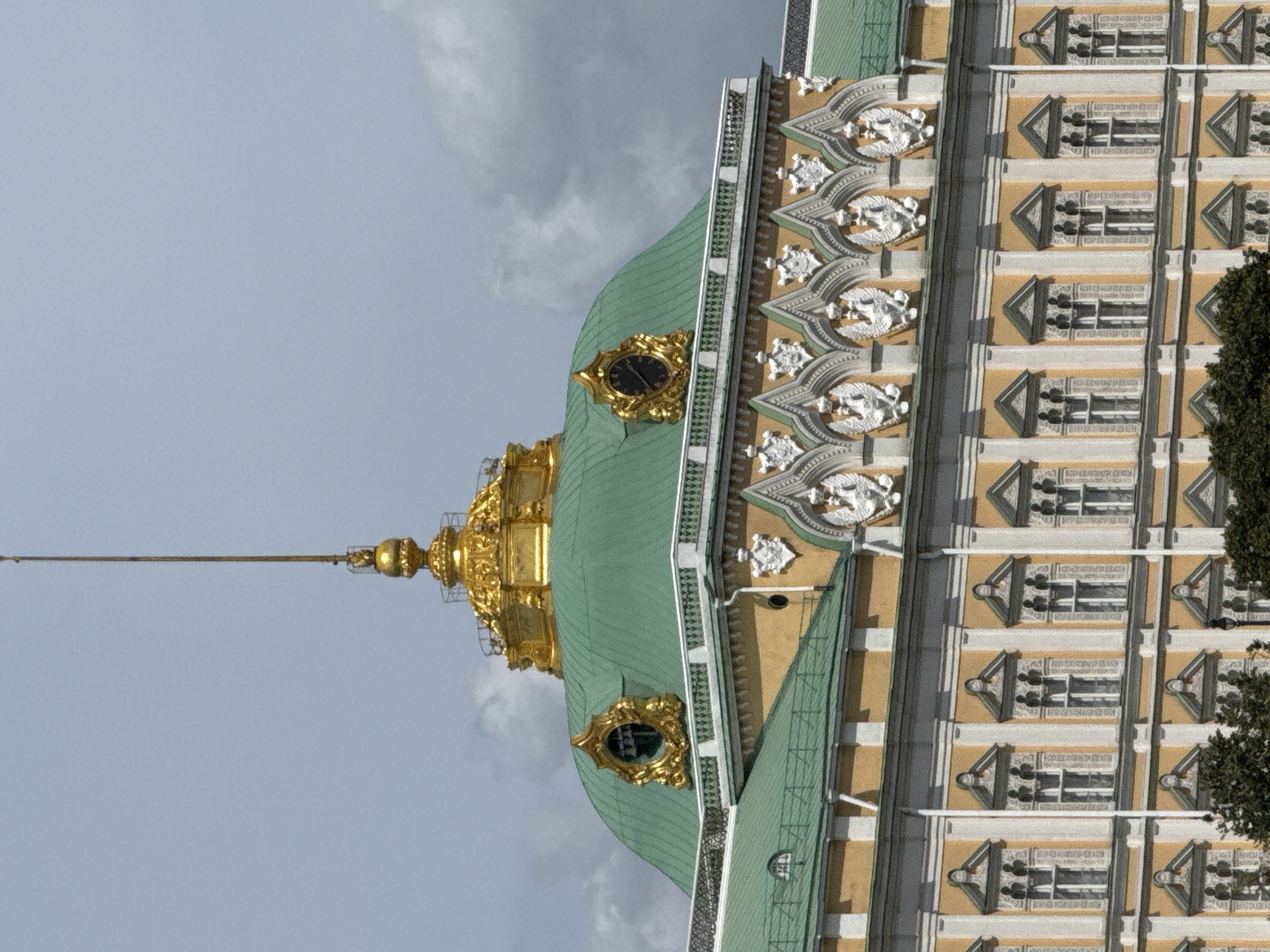 The image displays a vertical view of an ornate, historical building, likely a palace or cathedral, with a prominent green, domed roof and gilded architectural elements. The building is characterized by intricate detailing, including decorative carvings or sculptures adorning its facade, and a series of windows with elaborate surrounds. A tall, golden spire extends upwards from the roof. The sky above is a pale blue with scattered clouds, suggesting it is daytime, possibly late morning or afternoon.
The perspective is from below, looking up towards the top of the structure. The building's yellow facade is divided into sections by white vertical trim, and each section features multiple repeating window designs. Some windows are framed by triangular pediments and appear to have multiple panes. The green dome is partially visible, and a golden, ornate clock face is embedded within it, surrounded by decorative gold trim. Another smaller, circular window with gold framing is visible lower down on the dome. The overall impression is one of grandeur and historical significance. The scene appears to be set in a well-maintained, possibly park-like area, with a dark green tree visible on the right edge of the frame. There is no text visible in the image. The lighting suggests a slightly overcast or hazy day, with soft shadows.