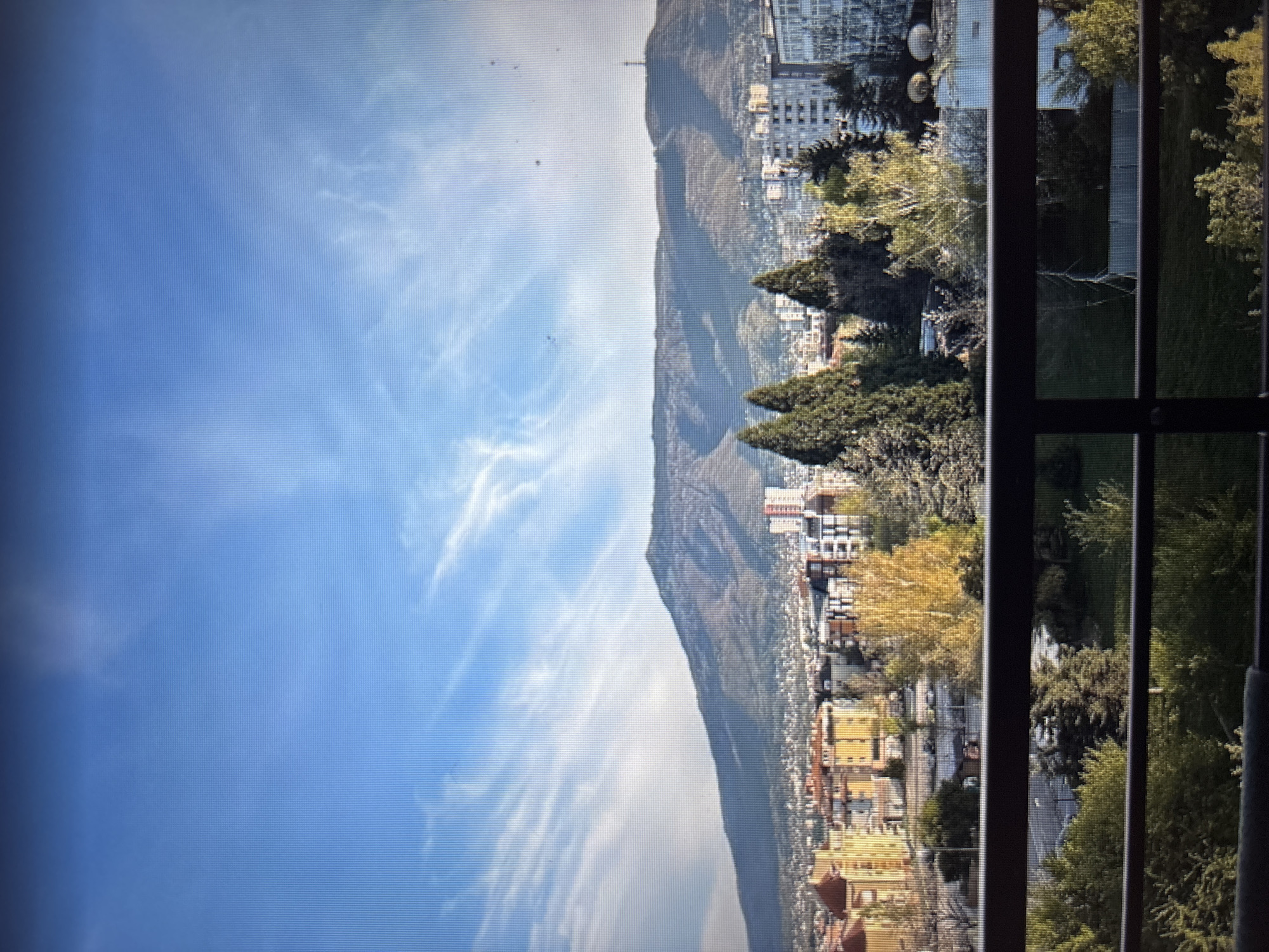 The image captures a view of the Vitosha mountain range from Sofia, Bulgaria. In the foreground, a dark railing of a balcony or terrace is visible, with lush green trees and foliage beneath it. Beyond the greenery, a cityscape unfolds, characterized by a mix of residential buildings of varying architectural styles and heights. Prominent among these are several multi-story apartment blocks, some with light-colored facades and others with orange or yellow hues. Several tall coniferous trees stand out amongst the buildings, contributing to the urban landscape.
The mid-ground is dominated by the imposing presence of Vitosha mountain, a large, rugged mountain with a barren, rocky peak. The mountain slopes show signs of vegetation and are cast in shadow in some areas, suggesting a late afternoon or early morning light. The sky above is a vibrant blue, streaked with wispy white clouds that add depth and texture to the scene. A small tower or antenna is visible on a ridge of the mountain in the distance. The overall impression is one of a city nestled against a majestic natural backdrop under a clear sky.