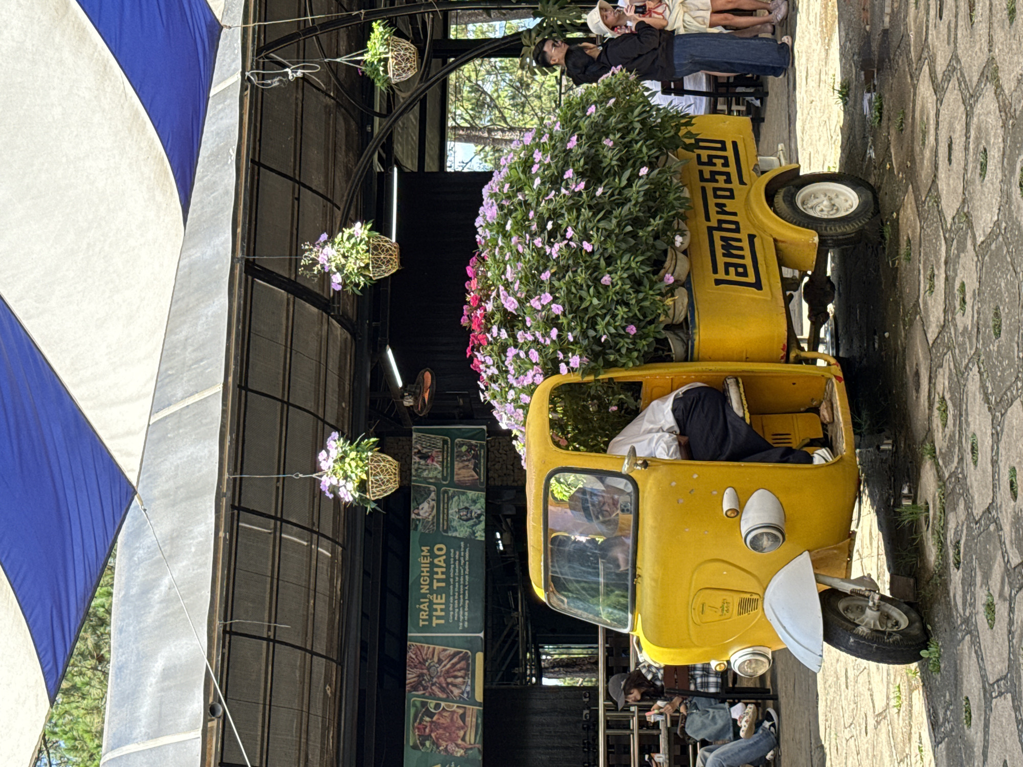 The image shows a yellow three-wheeled vehicle with the word "lambro550" written on the side. The vehicle is filled with a large bouquet of purple and pink flowers. It appears to be parked at a restaurant or tourist spot in Da Lat, Vietnam.
A person is seated inside the vehicle. A sign with the words "TRẢI NGHIỆM THỂ THAO" is visible in the background. The environment suggests an outdoor setting with a paved surface. Other people can be seen standing near the vehicle and seated at nearby tables.