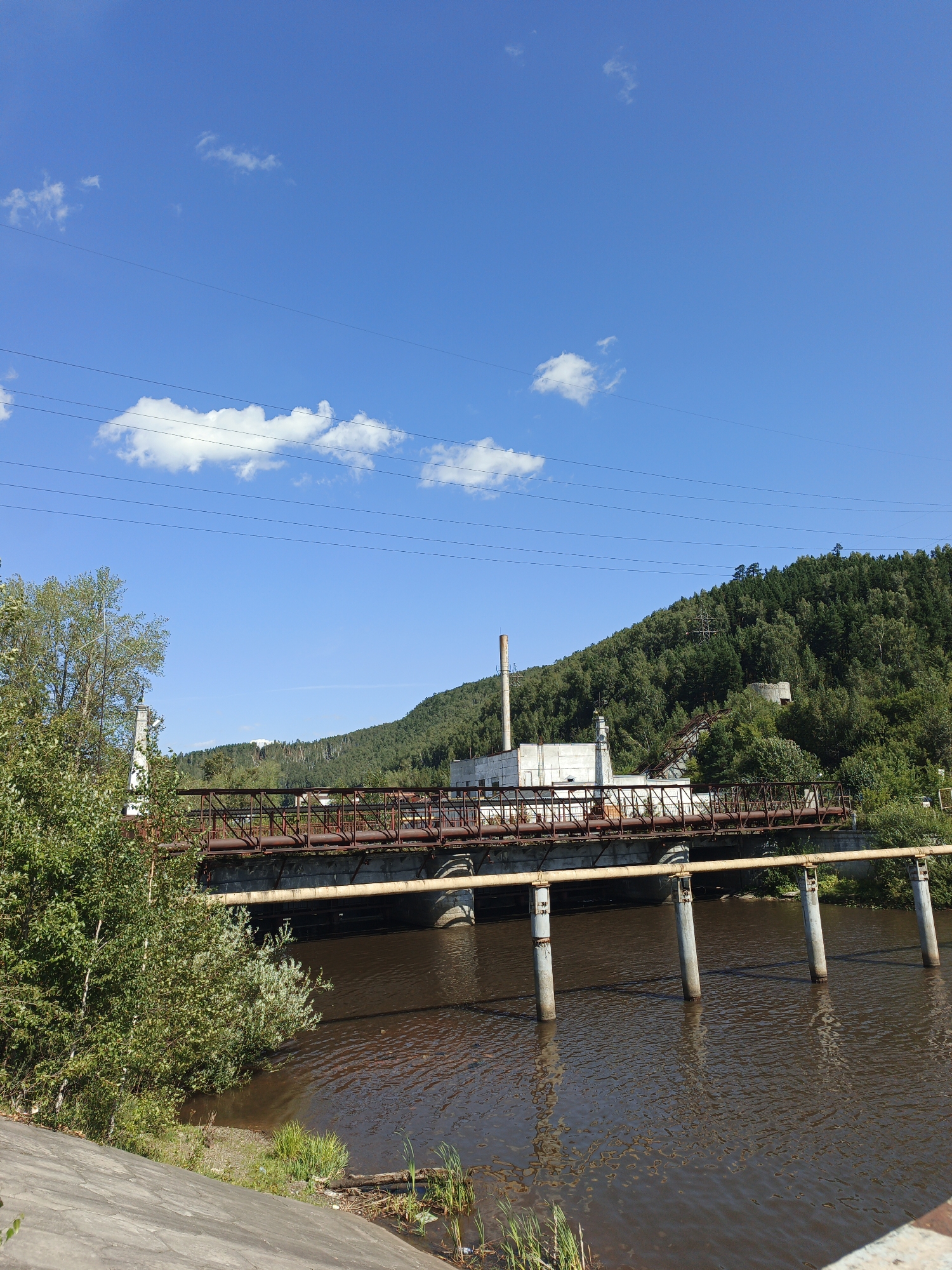 This is a photograph taken on a sunny day, looking across a body of water towards a bridge and an industrial structure. The sky is bright blue with scattered white clouds and several parallel power lines are visible. The water is dark brown and appears to be a river or lake. On the left bank, there is a concrete embankment and dense green foliage.
The bridge is constructed of weathered, rusty metal and spans the water. It carries a wide yellow pipe supported by concrete pillars. Behind the bridge, an industrial building complex is situated against a densely wooded hillside. This complex features a tall, cylindrical smokestack and several smaller buildings. The hillside is covered in lush green trees.
The specific location in the city of Zlatoust, Russia, is the Ufaleysky Highway bridge over the Ai River, adjacent to the Zlatoust Thermal Power Plant.