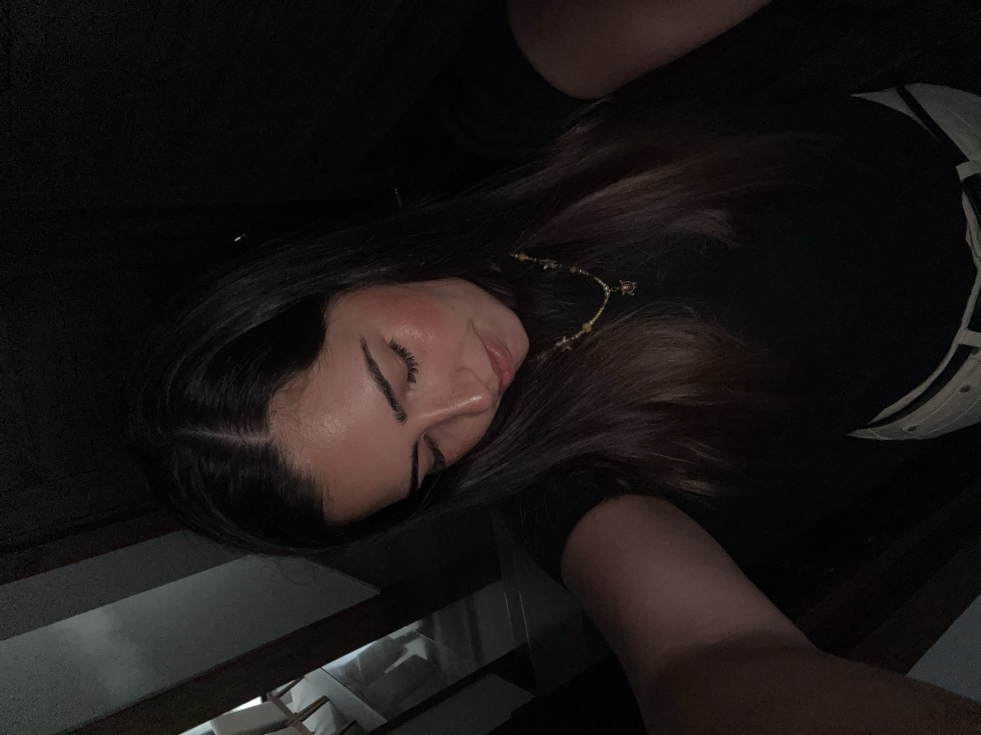 A young woman with long brown hair lies on a dark wooden surface, possibly a floor or stairs. She is wearing a black top and a white belt. Her eyes are closed, and she has on makeup. A beaded earring or hair accessory is visible near her face. The lighting is dim, making it difficult to discern further details. The image does not provide any information to place it in San Bernardino Tlaxcalancingo, Mexico.