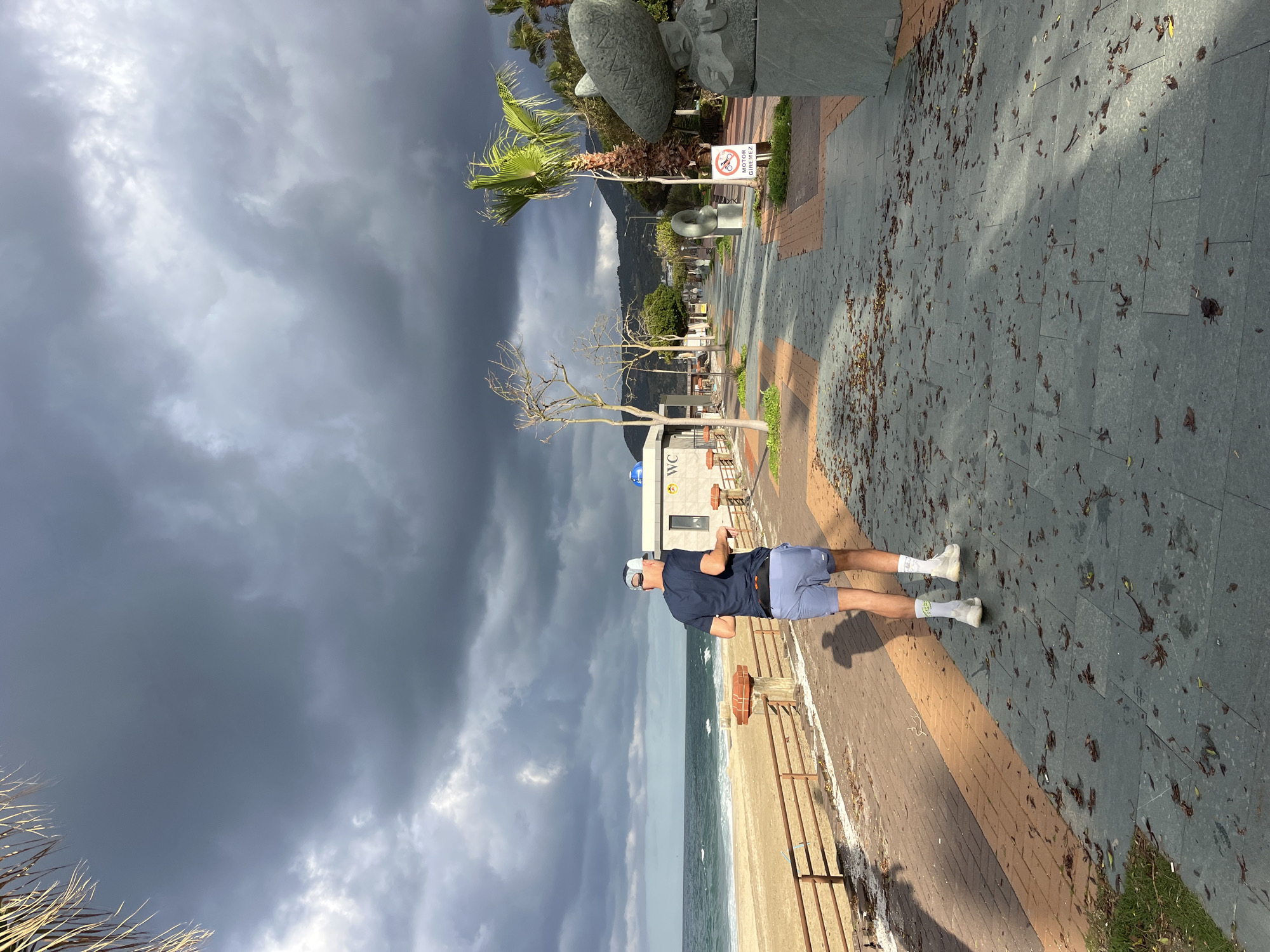 A middle-aged man wearing a navy t-shirt, light blue shorts, white socks, and a white cap stands on a paved walkway along the beach in Alanya, Turkey. The beach is to his left, with a metal railing separating the walkway from the sand. The sea is visible in the background, under a dramatic sky filled with dark storm clouds.
Behind the man, across the walkway, is a restroom (WC) building. A row of bare trees and a palm tree line the sidewalk further down. Sculptures are visible along the walkway in the distance, along with a sign with a red circle and diagonal line, indicating a restriction of some kind.
The walkway is paved with grey tiles interspersed with sections of reddish-brown bricks. Fallen leaves are scattered across the walkway. The lighting suggests late afternoon, with strong shadows cast by the figure and objects. A mountain range is faintly visible in the far distance.