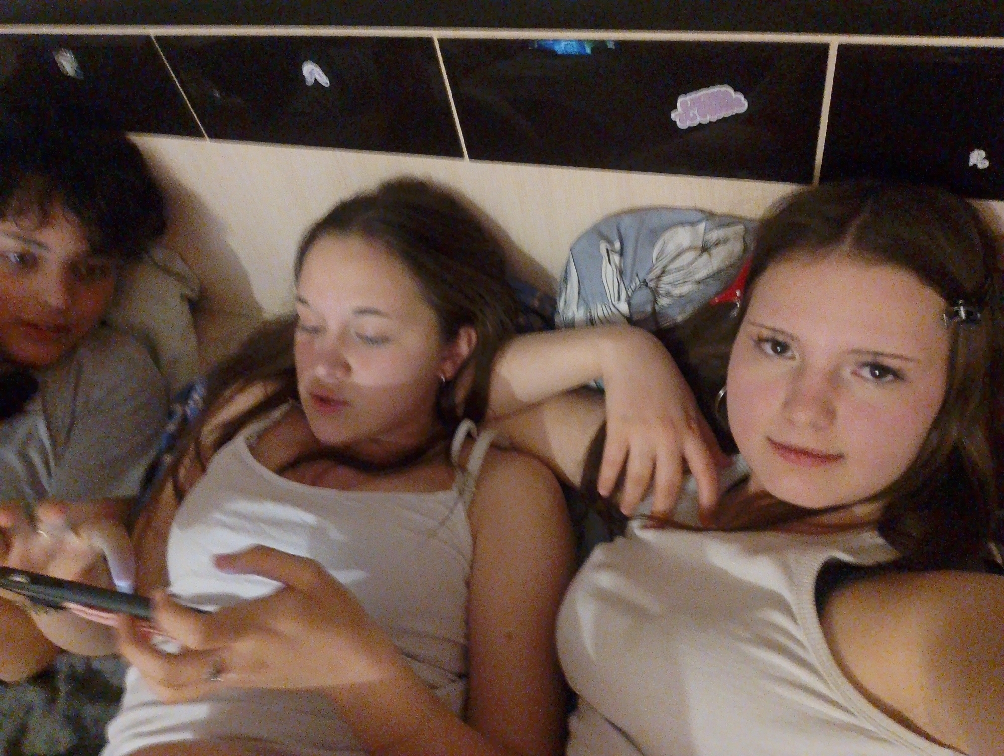 Three individuals are indoors in what appears to be a domestic setting, likely a bedroom. Two young women are lying down side by side; one is looking directly at the camera with a slight smile, while the other has her eyes closed. A third individual, with short dark hair and wearing a grey t-shirt, is partially visible on the left side of the frame, holding a smartphone and looking at it. All three are casually dressed, wearing tank tops. The background consists of a headboard with decorative elements like white and purple stickers on a black background. The location is most likely inside a house in Фоки, Russia.