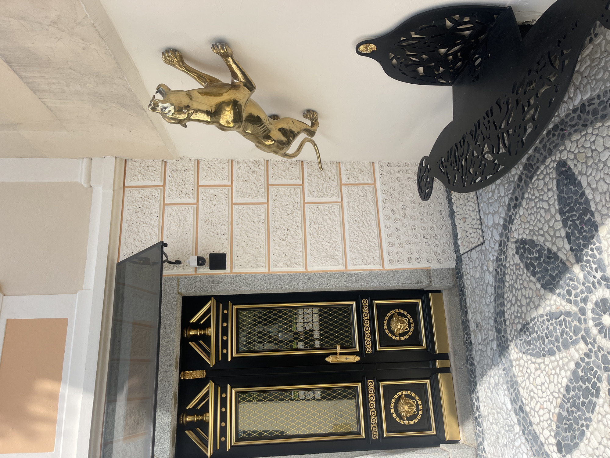 The image shows an ornate black door with gold detailing, including Medusa heads, on the facade of a building. To the right of the door, a golden sculpture of a panther is mounted on the wall, appearing to be climbing. In the foreground, a black metal bench with intricate cut-out designs is visible, positioned on a cobblestone pathway with a mosaic pattern. The wall next to the door is textured and features a row of light-colored blocks. A security camera is mounted on the wall above the door. The overall scene suggests a sophisticated entrance, possibly at a hotel or upscale establishment, located in the historic center of Brixen (Bressanone), Italy.