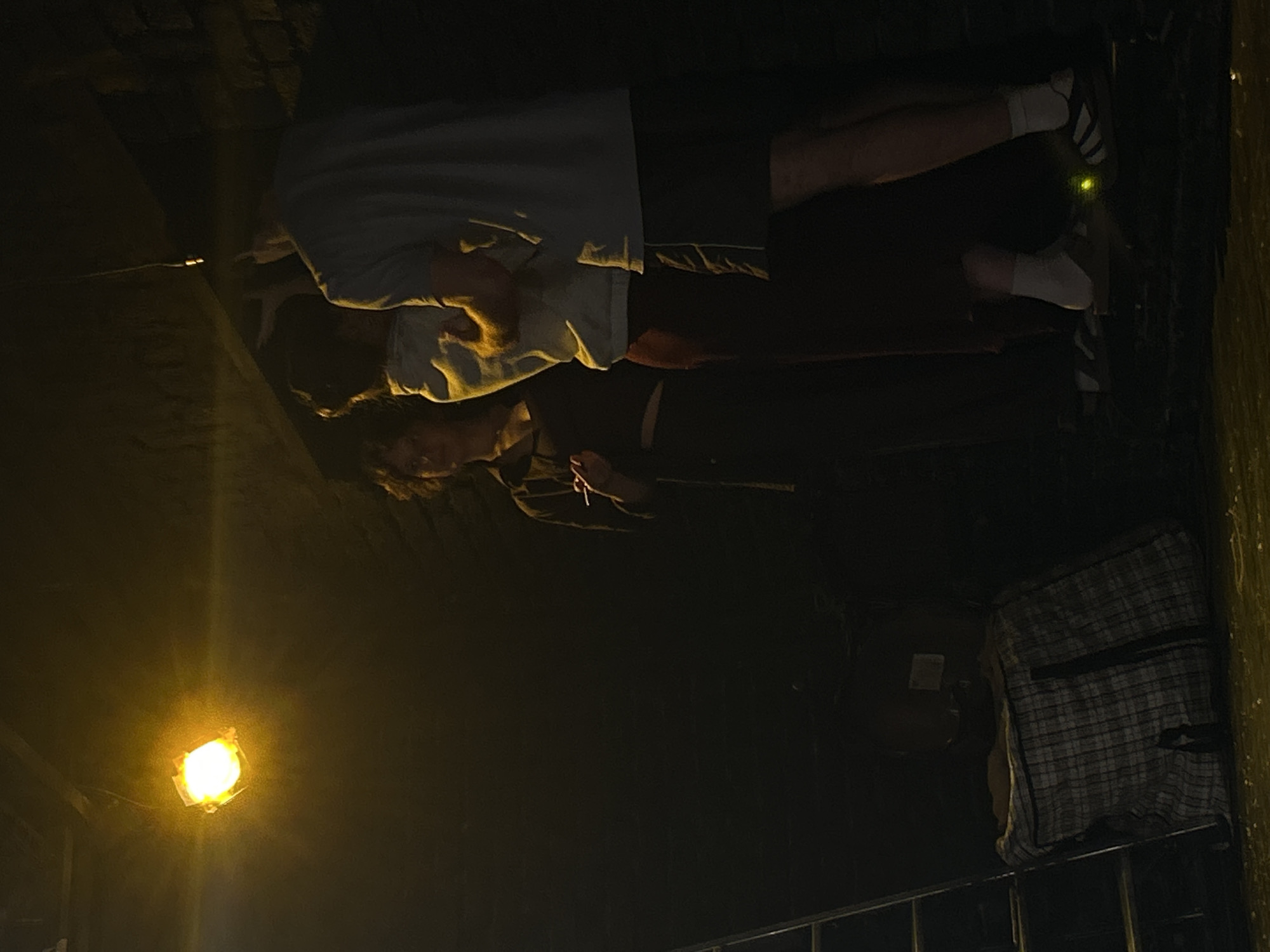 A group of three individuals are standing in a dimly lit, indoor space, possibly a basement or storage area, in Unknown, Unknown. A bright yellow light source hangs from the ceiling, illuminating the scene. The person on the left, a woman, is wearing dark clothing and appears to be holding a cigarette. To her right, a man in a light-colored hoodie and dark pants is facing away from the camera, with his arm around another person who is partially obscured. The third individual, a man on the far right, is wearing a light blue t-shirt and dark shorts. The floor is covered with a plaid duffel bag and a ladder is visible on the left side of the frame. The walls appear to be constructed of brick.