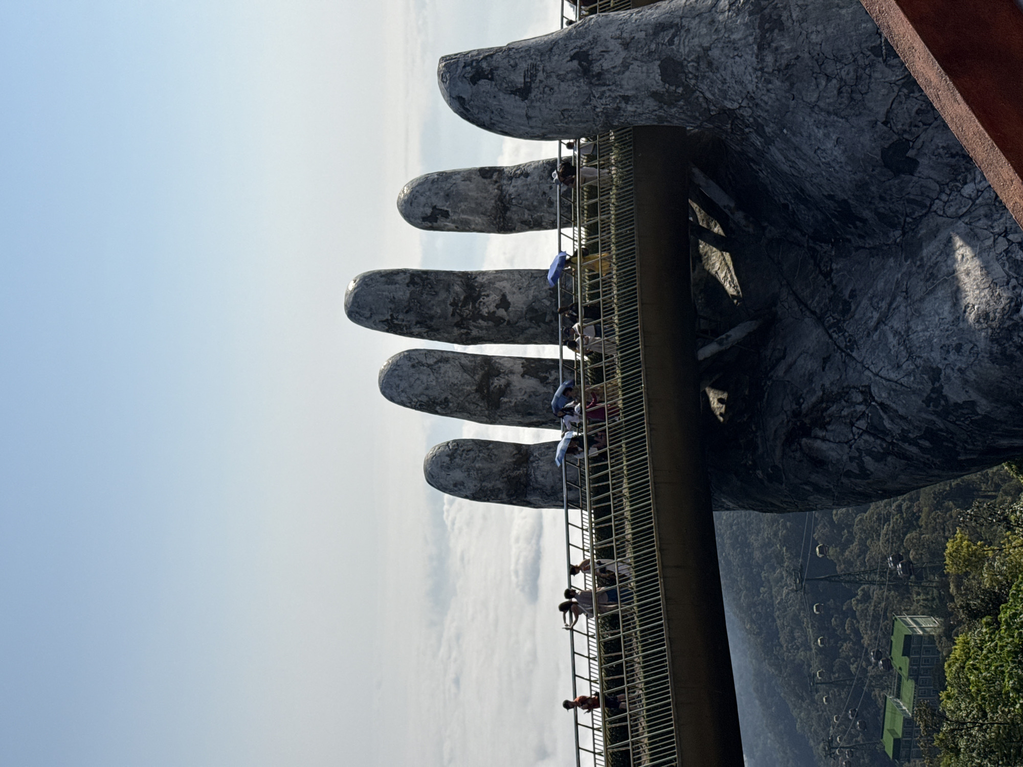 The image captures the Golden Bridge (Cầu Vàng) at Bà Nà Hills, Đà Nẵng, Vietnam, situated at a high elevation. A curved, golden-colored pedestrian bridge, featuring white metal guardrails, traverses the mid-ground. It is structurally supported by two colossal, weathered gray concrete hands, designed to appear like ancient stone; one hand is prominent on the right, displaying five finger-like structures, while parts of the second hand are visible on the left. Approximately 20-30 individuals, identified as tourists, are observed walking along the bridge in various directions. Several individuals are holding blue or light-colored umbrellas. One person on the left side of the bridge is actively taking a photograph with a mobile device. The sky above the bridge is clear and light blue. Below the bridge, a thick stratum of white clouds partially obscures the lower mountain landscape. Further beneath the clouds, lush green forested mountains are visible, with a prominent cable car system featuring multiple dark-colored gondolas traversing the slopes. Two multi-story buildings with distinct green roofs are visible nestled within the lower forested terrain.