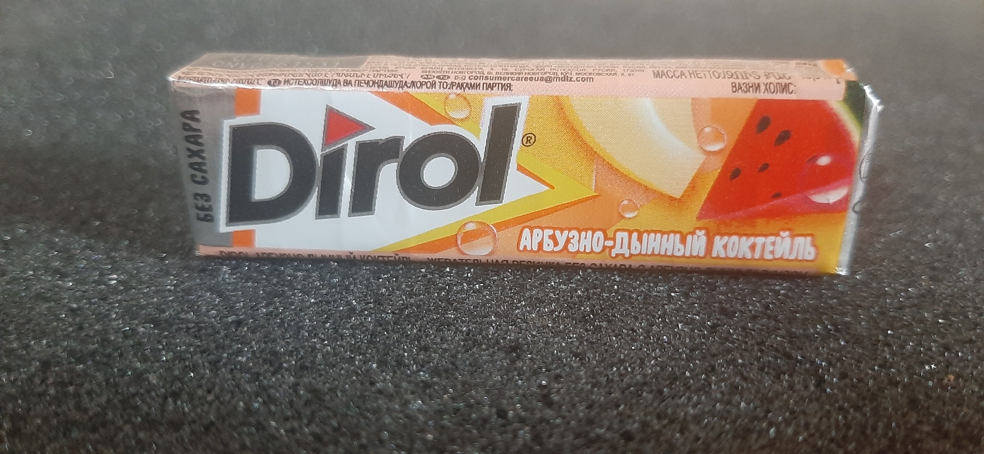 A pack of Dirol chewing gum, specifically the "Watermelon-Melon Cocktail" flavor, is shown on a dark, textured surface. The packaging is predominantly white, with orange and yellow accents. The Dirol logo is prominent in black. The flavor is written in Russian as "АРБУЗНО-ДЫННЫЙ КОКТЕЙЛЬ". The gum is labeled "БЕЗ САХАРА" (sugar-free). The image is taken from a slightly elevated angle, focusing on the product. There is no discernible location in the city of Yessentukskaya, Russia, visible in the image.