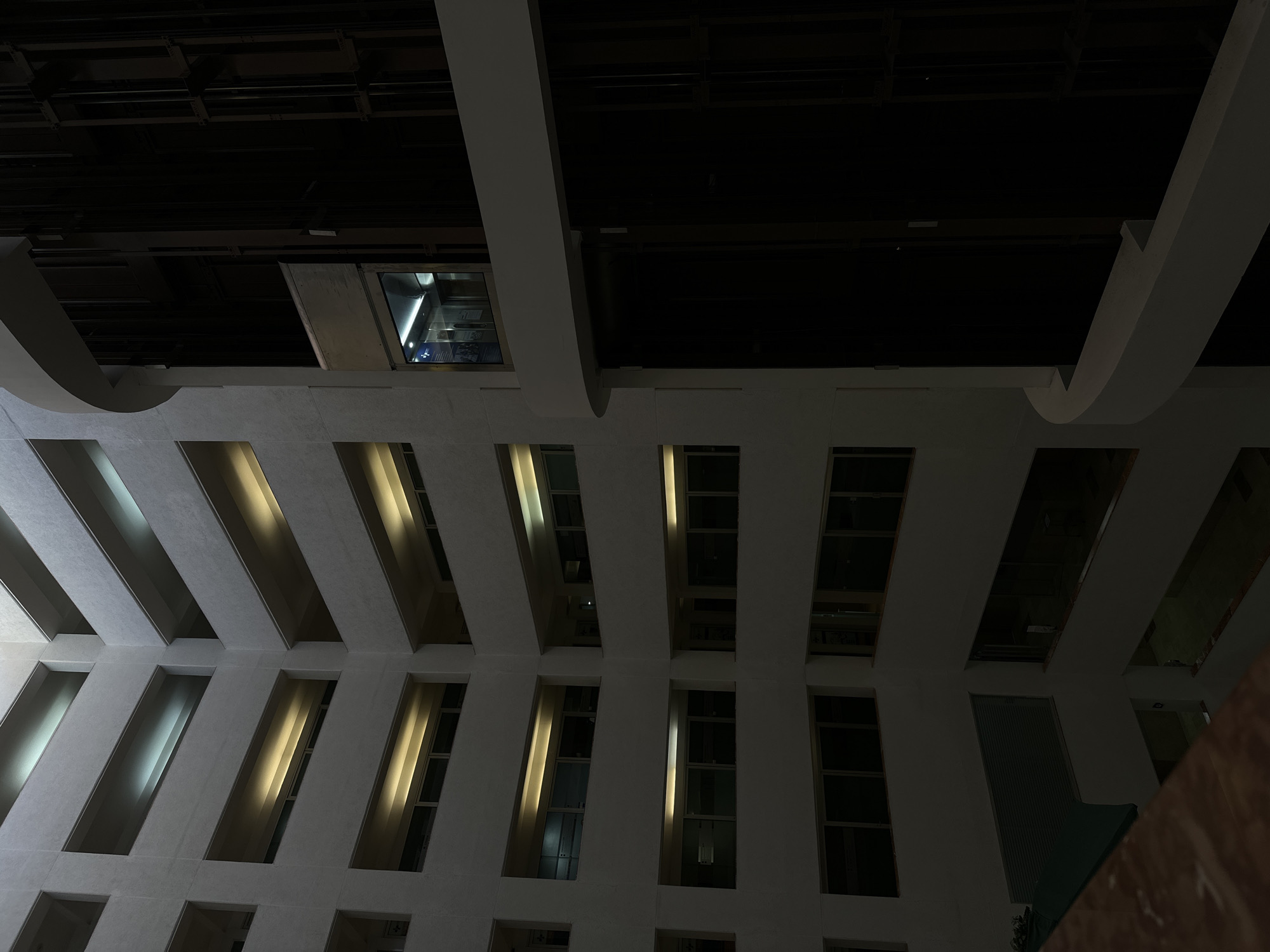 The image presents an upward-looking view within a modern building's interior, likely an atrium or a multi-story hall. The dominant feature is a repetitive pattern of rectangular, deeply recessed openings or windows on a light-colored, possibly white or light-grey, wall. Many of these openings glow with artificial light, some emitting a warm, golden hue, while others display a cooler white light.
The upper portion of the scene is significantly darker, revealing an industrial-style ceiling with visible dark-colored structural beams, pipes, and other infrastructure. Curved, light-colored architectural elements extend across the top of the frame, creating visual breaks.
Towards the upper left, a glass-encased elevator cabin is visible within an opening in the dark ceiling. The elevator's interior is brightly lit, and a blue sign with unreadable text can be vaguely seen inside.
The overall impression suggests an evening or nighttime setting, as artificial lighting is prominent. No people or specific activities are depicted. The architecture is characterized by strong vertical lines, repetitive forms, and a stark contrast between illuminated and shadowed areas.