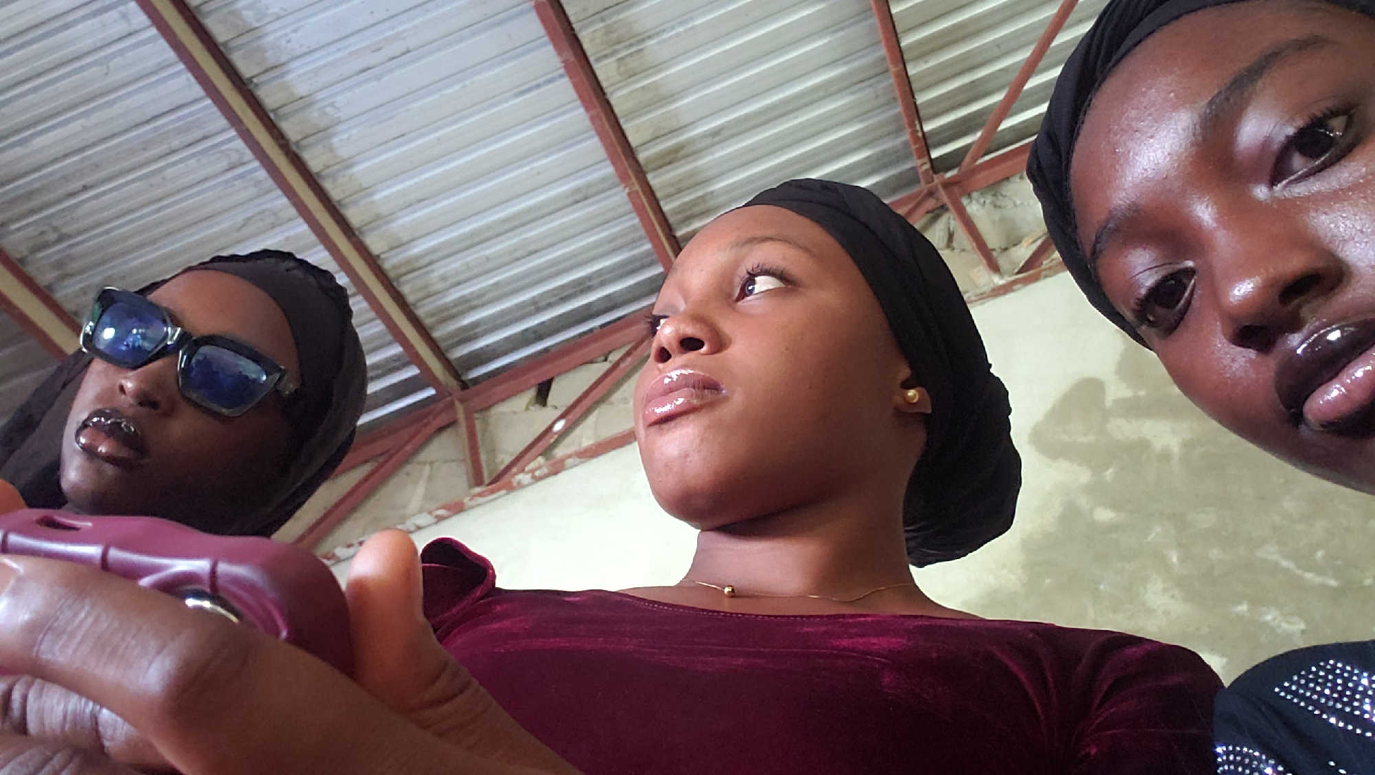 Three individuals, presumed to be young women, are depicted from a low-angle perspective, looking upwards toward the camera. The setting is an indoor or semi-sheltered area in Narayi, Nigeria, characterized by a corrugated metal roof supported by red-painted metal trusses and beams, with a textured, light-colored wall in the background.
The individual on the far left wears dark, rectangular sunglasses with blue-tinted lenses and a black head covering. Their lips are glossy, and a metallic oral appliance or jewelry is visible on their lower teeth. They are holding a purplish-pink electronic device, possibly a smartphone, with their right hand.
The central individual is seen from the chest up, wearing a black head covering, a dark red long-sleeved garment, a single gold stud earring in their right ear, and a thin gold chain necklace with a small spherical pendant. Their lips are glossy, and they are looking upwards and slightly to the right.
The individual on the far right is depicted in a close-up, showing their face from the nose upwards. They are wearing a black head covering, and their skin exhibits a high sheen. Their glossy lips are parted, with the tongue slightly protruding. A black garment with numerous small, reflective embellishments is visible on their upper chest/shoulder.