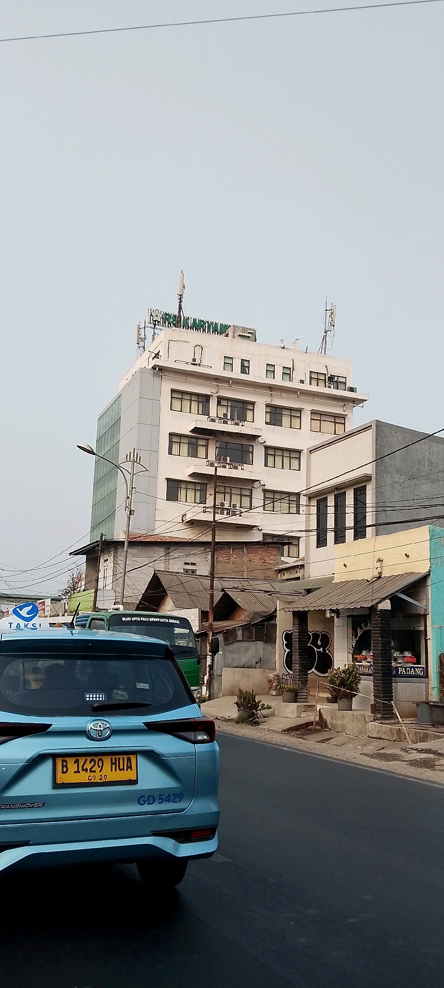 A light blue car, possibly a Toyota, is parked on an asphalt road in Bekasi, Indonesia. The car has a yellow license plate with the number B 1429 HUA. Behind the car, there is a tall white building with multiple floors and windows. The building has signage at the top that reads "RIAN KARYAMAN". To the right of the white building, there are smaller structures, including one with a sign that says "PADANG MASAKAN". The sky is overcast, and there are several utility poles and wires visible. A green truck is also present on the road, partially obscured by the blue car. The overall scene depicts a street view in an urban environment.