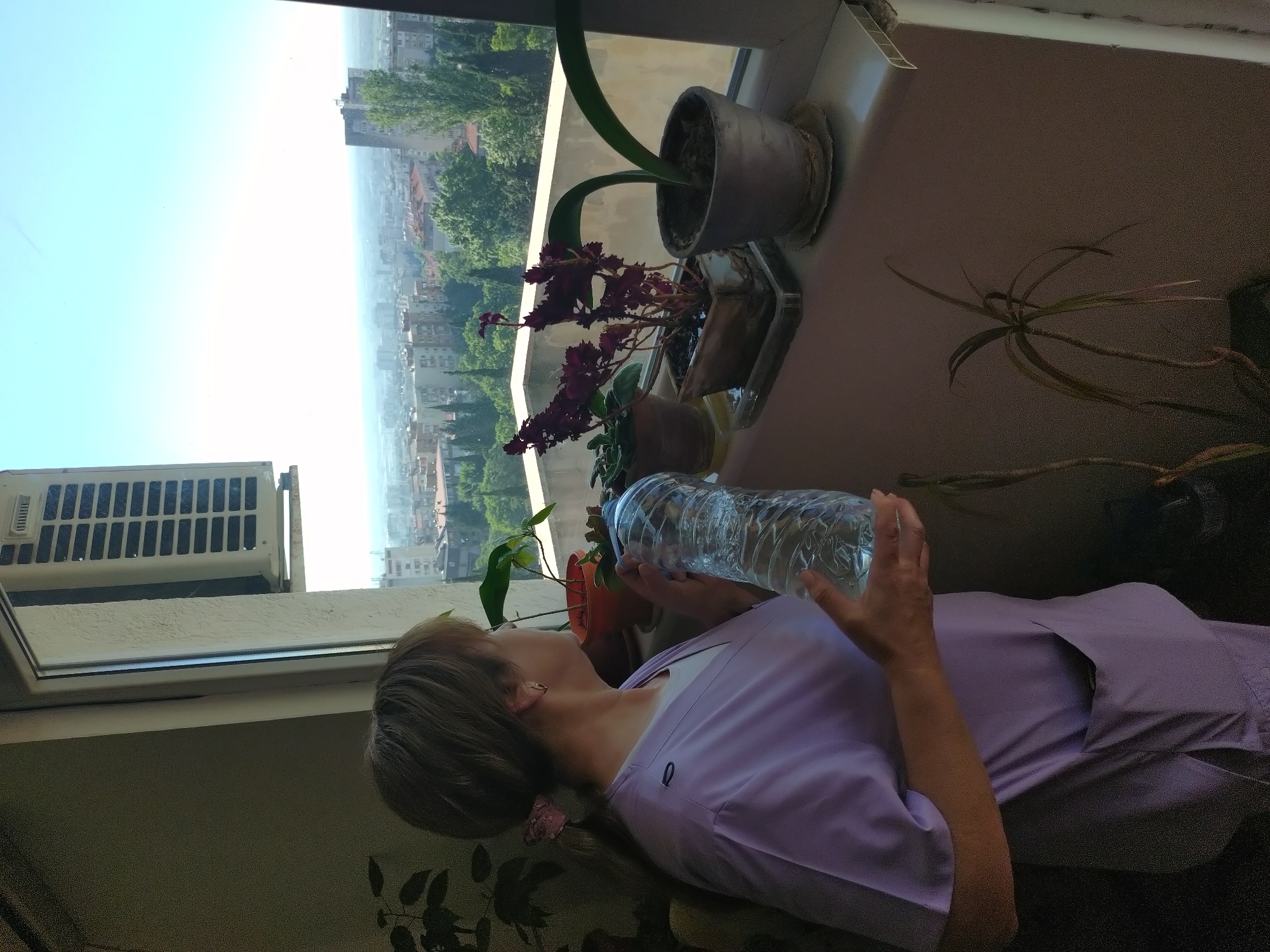 A woman in a light purple medical scrub top holds a clear plastic water bottle in her hands. She is standing near a window with several potted plants on the windowsill, including a plant with purple flowers. Outside the window, a city landscape is visible under a bright, clear sky. An air conditioner is visible in the corner of the window. The location is Stara Zagora, Bulgaria.