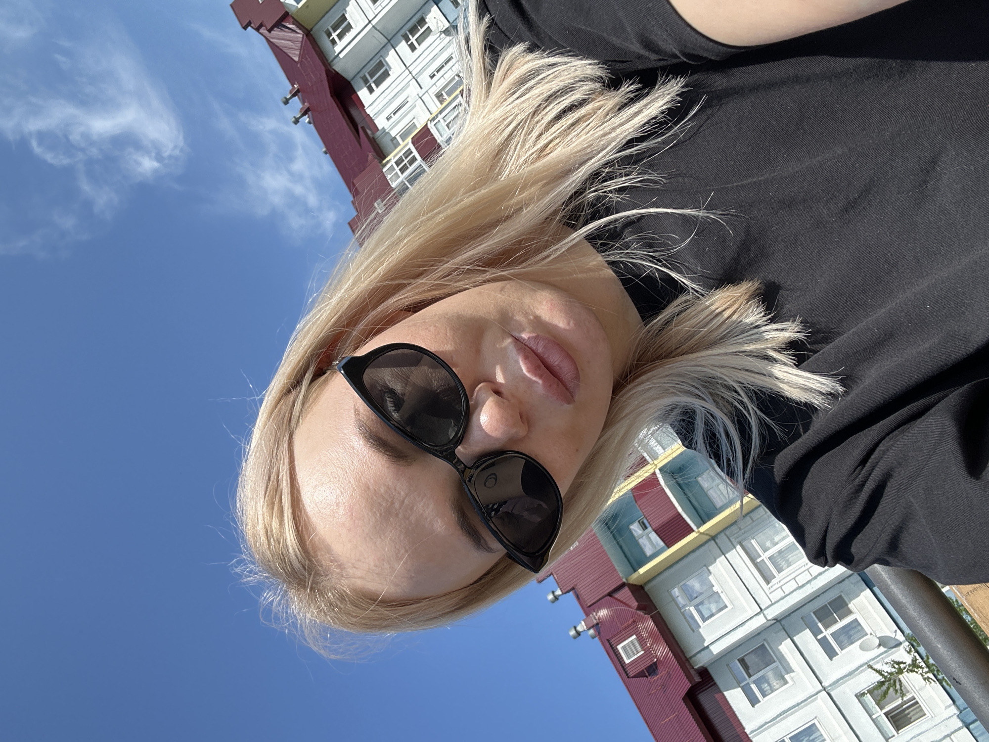 A woman with blonde hair and sunglasses takes a selfie outdoors. She is wearing a black t-shirt. In the background, a multi-story building with a red roof and light blue and yellow siding is visible against a clear blue sky with scattered clouds. The camera angle is upward.