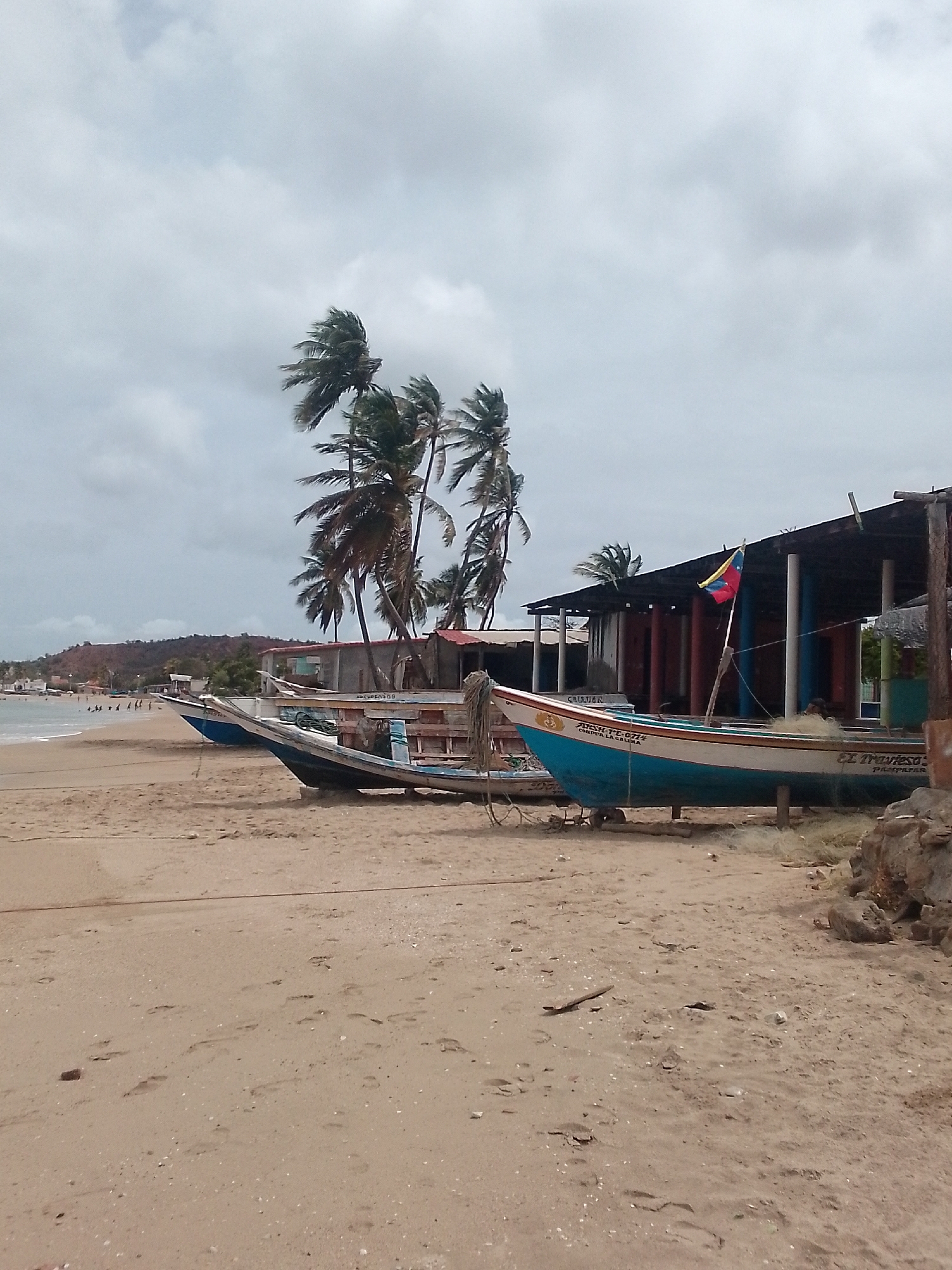 The scene is a beach in Juan Griego, Venezuela. In the foreground is sand, marked with numerous footprints. Three boats rest on the beach, ranging in color from blue to light blue. The boats are marked with text in Spanish. Behind the boats, a simple wooden structure with a roof is visible, supported by columns, and a Venezuelan flag flies from it. Palm trees sway in the background, suggesting a windy day. In the distance, people are visible on the beach near the water's edge, and a hillside is visible in the far background. The sky is overcast with heavy clouds.