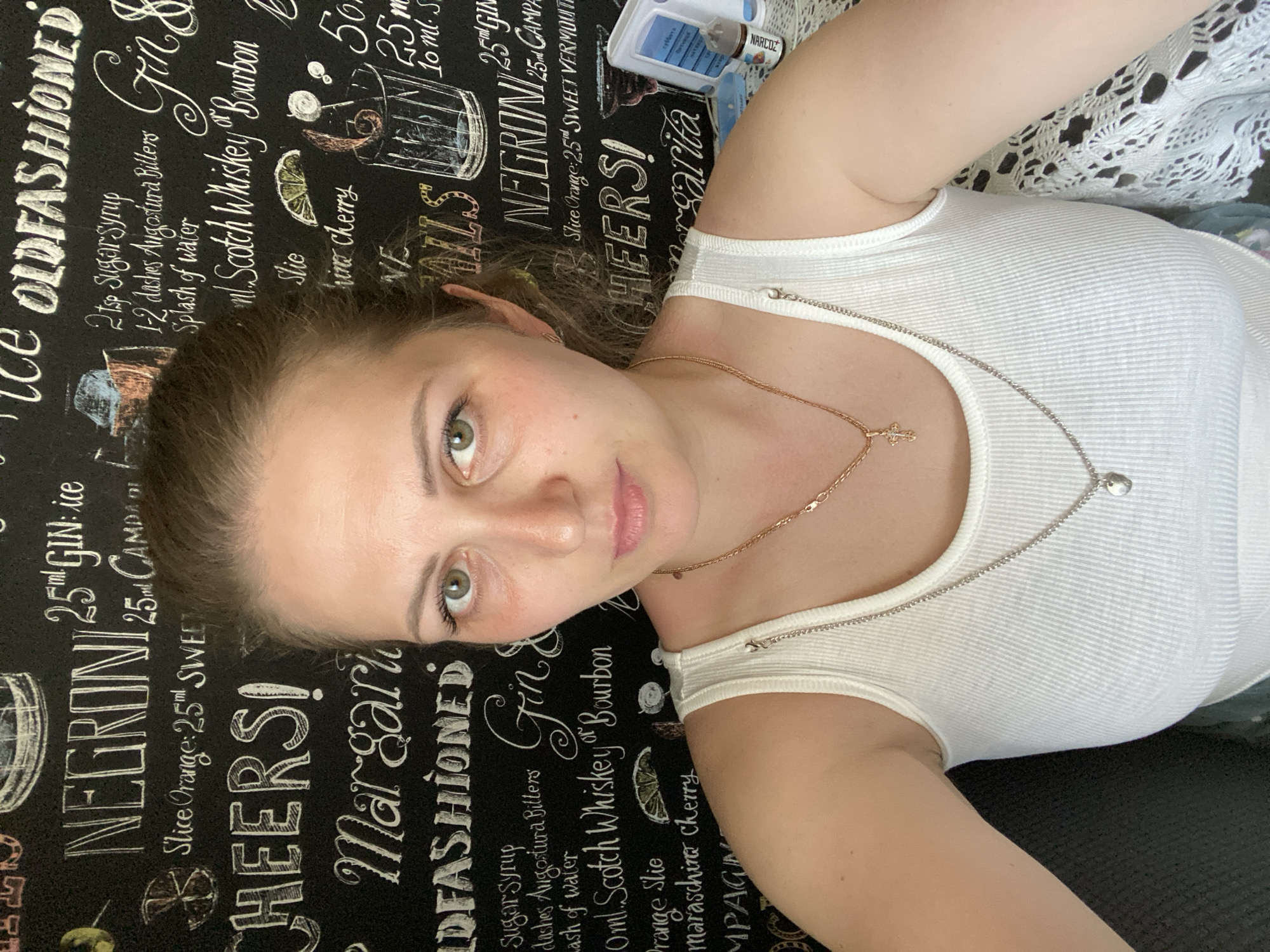 A woman lies at an angle against a black surface printed with cocktail recipes and names, including "NEGRONI," "CHEERS!," "Margarita," and "OLD FASHIONED" in white text and decorative illustrations. She wears a white ribbed tank top and two necklaces, one silver and one gold. A white and blue plastic container labeled "NARCOZ" sits to the upper right of the woman. The location cannot be confirmed as Марковский, Russia, based on provided image data.