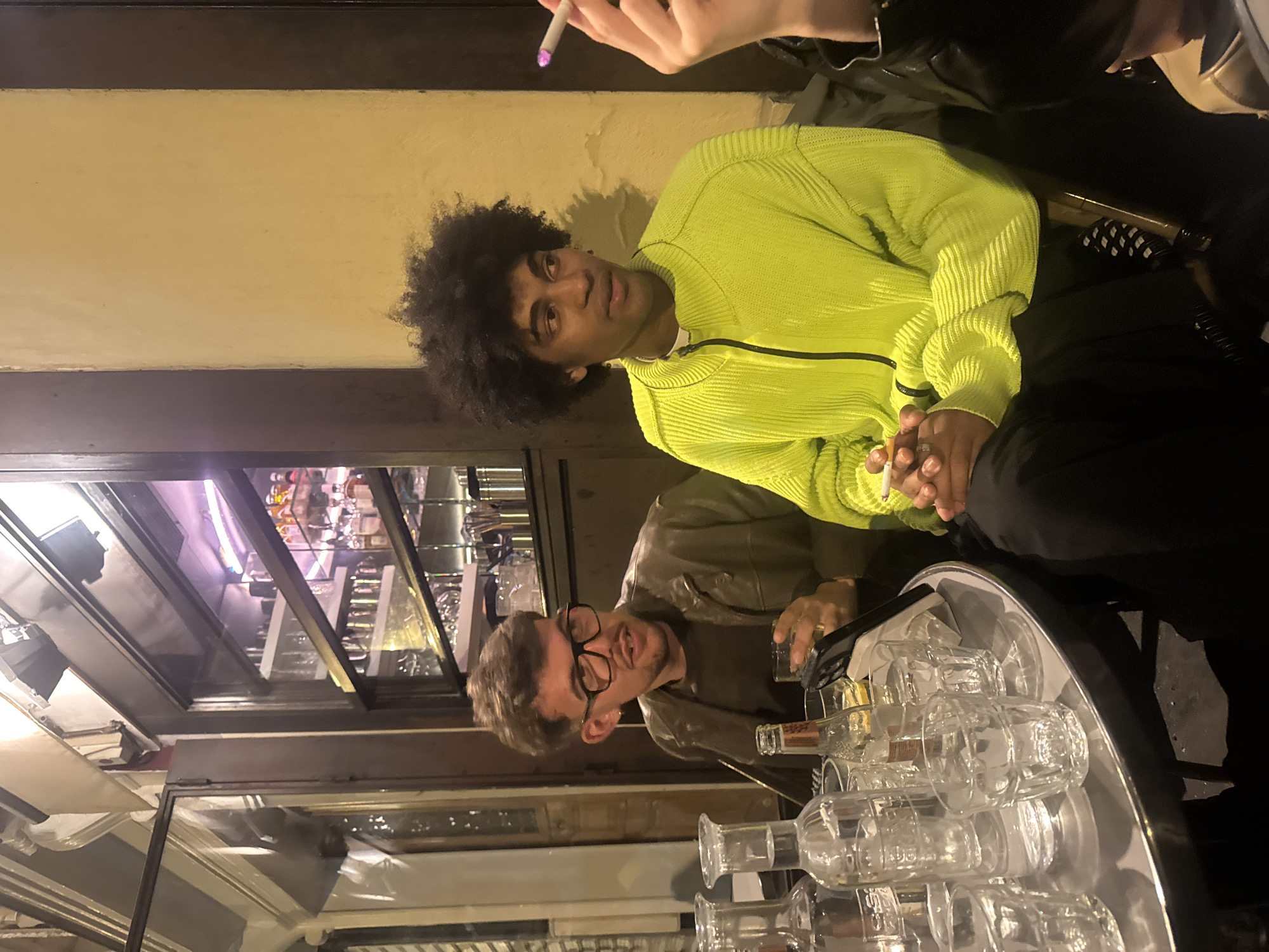 The image captures a casual social scene, likely in a bar or restaurant in Paris, France, given the architectural style of the building and interior design. Two men are prominently featured. One is wearing a vibrant yellow-green ribbed jacket, has dark curly hair, and is holding a cigarette. The other man, wearing glasses and a brown jacket, is seated at a table. There's a tray filled with empty glasses and liquor bottles on the table. Other individuals can be seen in the background, one holding a cigarette. The environment appears to be a partially enclosed area with glass windows, possibly an outdoor seating section, with the city visible through the glass.