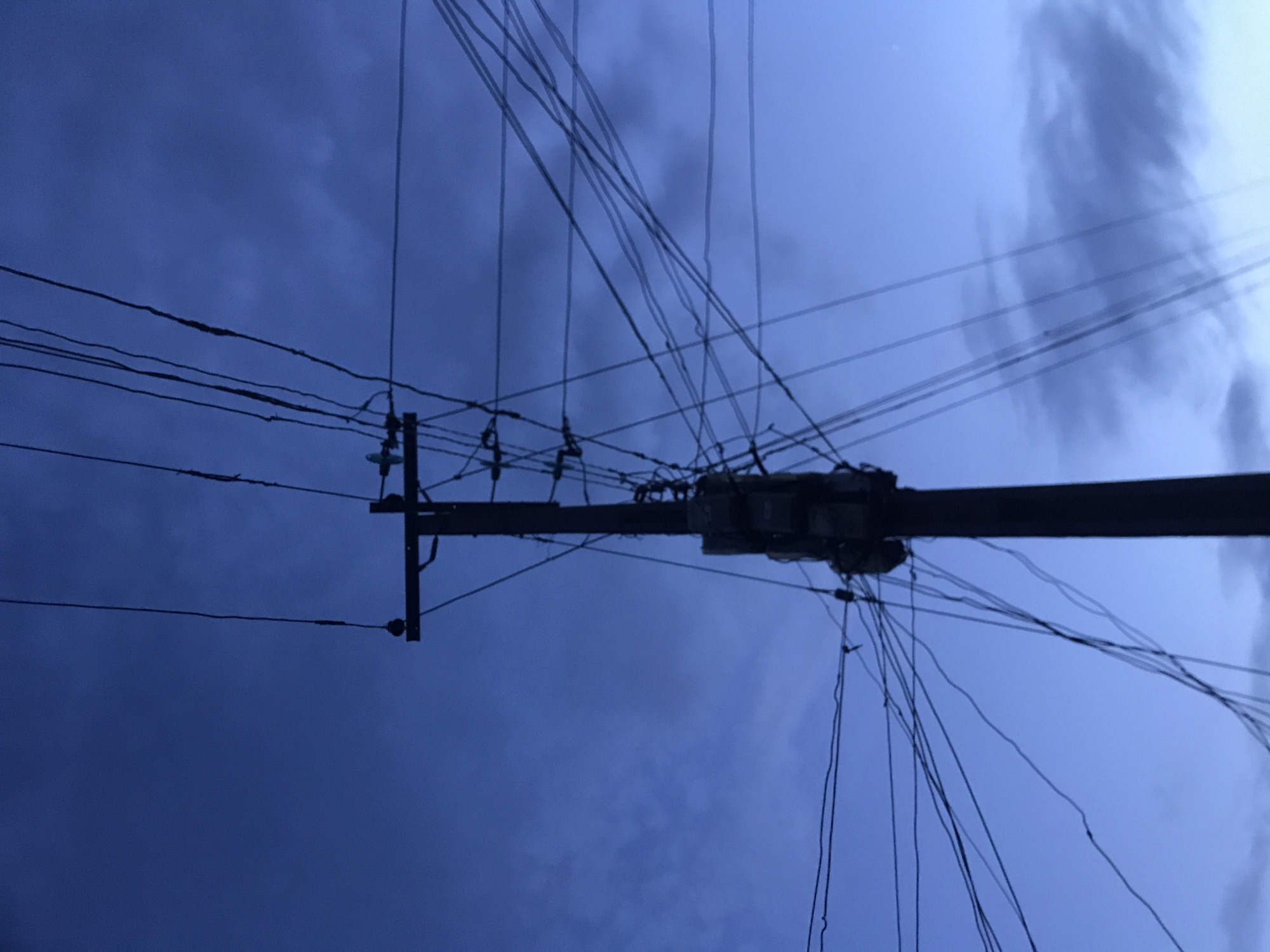The image depicts a utility pole and its overhead electrical infrastructure silhouetted against a deep indigo sky, characteristic of twilight, in Rafin Par, Nigeria. The dark, vertical utility pole is centrally positioned, extending from the bottom edge of the frame upward. It features a horizontal crossbar near its apex, supporting several insulated wires. A dense and complex network of numerous electrical cables emanates from various points on the pole, particularly from multiple box-shaped utility devices mounted below the crossbar, fanning out and crisscrossing across the frame. The sky background contains diffuse, dark cloud formations, notably in the upper-right and lower-right portions of the image. No individuals, vehicles, or additional specific objects are discernible.