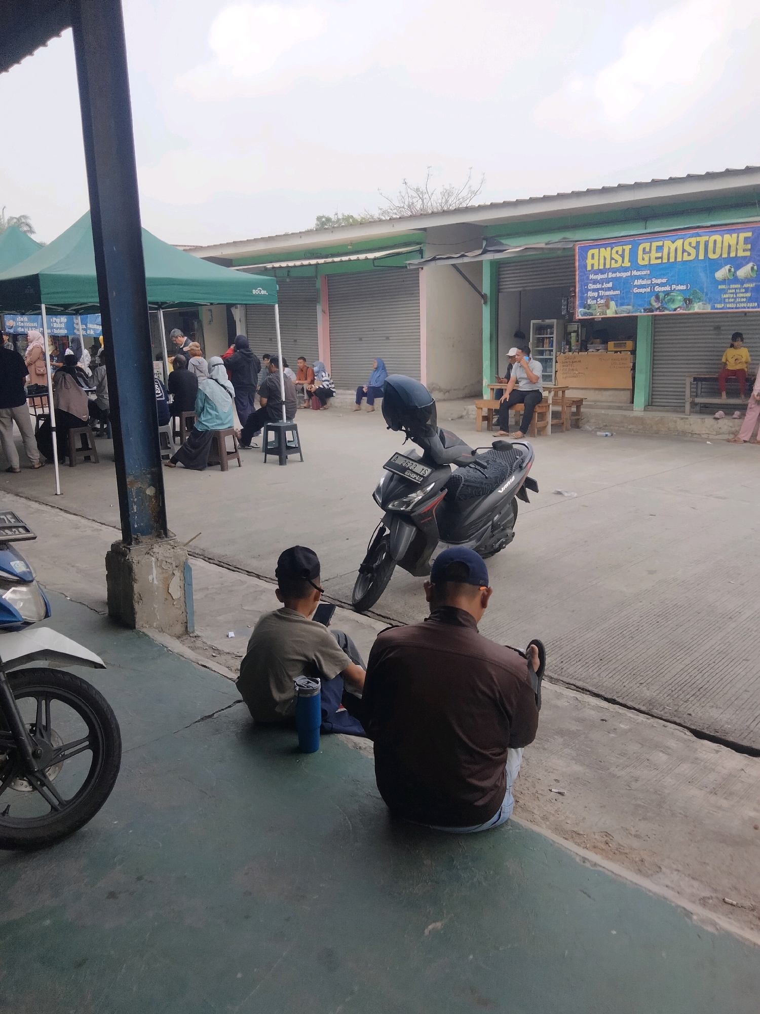 The image depicts an outdoor market scene in Sukaasih, Indonesia. In the foreground, two men are sitting on the ground, facing away from the camera. The man on the left wears a light brown t-shirt and blue jeans, and a blue tumbler sits beside him. The man on the right wears a dark brown jacket and blue jeans, and a black sandal is on his right foot. A black motorcycle is parked to the left of the foreground, with its front wheel visible.
In the mid-ground, a dark-colored motorcycle is parked. Beyond that, several stalls are set up under green tents. People are seen walking and sitting around these stalls. One stall has a blue banner with the text "ANSI GEMSTONE" and lists various items for sale. Further back, a building with shuttered windows lines the area. To the right, a young girl in a yellow shirt and pink pants sits on a wooden bench. The ground is paved with concrete and asphalt. The sky is overcast.