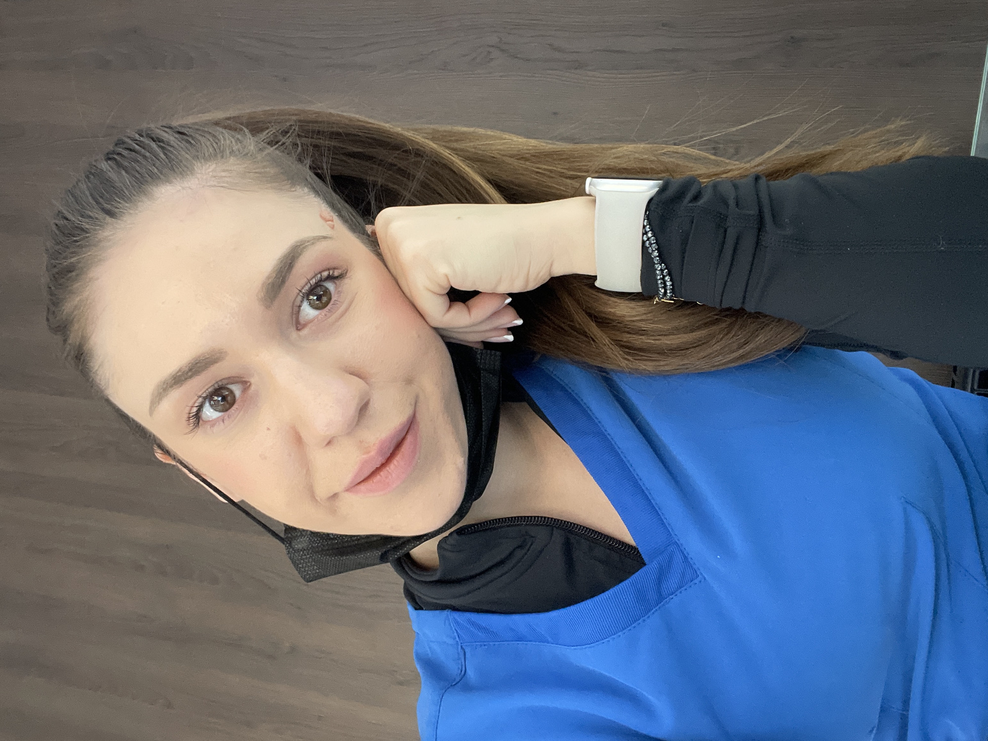A woman with light brown hair pulled back in a ponytail is looking at the camera. She has brown eyes, fair skin, and is wearing a black face mask pulled down under her chin. She is wearing a bright blue scrub top over a black undershirt and has an Apple Watch with a white band on her left wrist. Her hand is resting against her cheek. The background consists of a wood-grained surface. The picture appears to be taken indoors, possibly in a hospital or clinic. The image does not contain any information that could be used to determine the specific location in Sofia, Bulgaria.