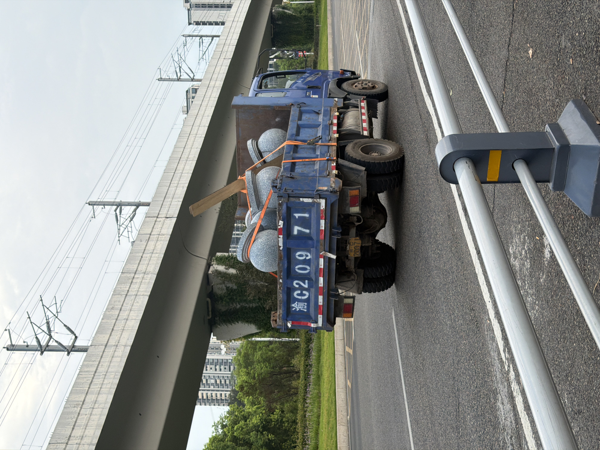 A blue flatbed truck is driving on a road in Jiangjin, China. The truck is carrying several large, round, grey objects that appear to be decorative stones or planters, secured with orange straps. A wooden plank is also visible in the truck bed. The truck has Chinese license plates that read "渝 C209 71". In the background, there is an elevated structure, possibly an overpass or a train track, with electrical wires. Buildings and trees are visible beyond the elevated structure. The sky is overcast. In the foreground, a metal guardrail is positioned along the roadside.