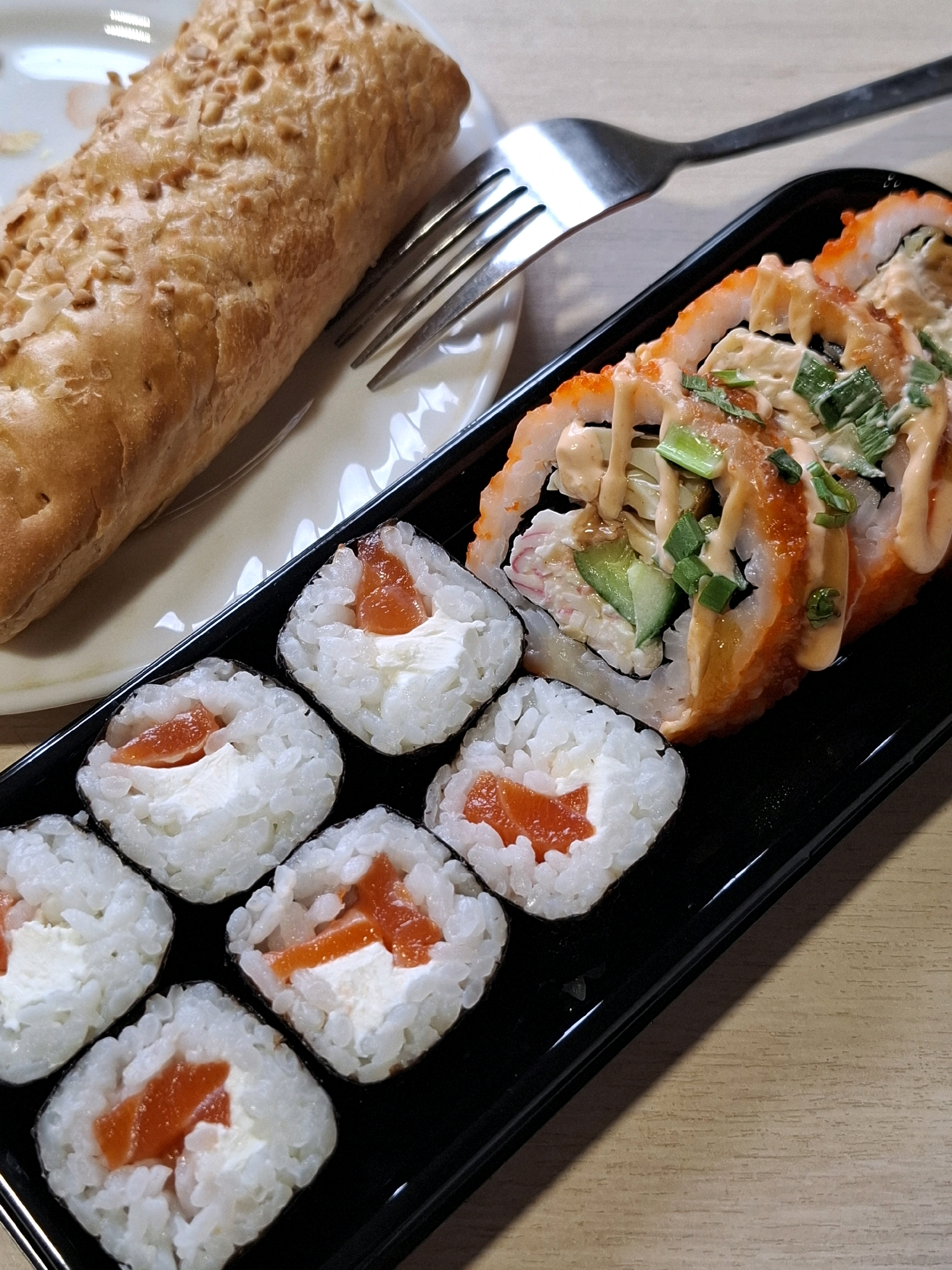 A partially visible baked good topped with sesame seeds rests on a white plate next to a metal fork. Adjacent to the plate, on a black rectangular platter, are several pieces of sushi. These include four maki rolls filled with rice, white cream cheese, and a piece of orange salmon, and one larger California roll topped with orange tobiko, creamy sauce, and chopped green onions. The scene is set on a wooden surface. The image depicts food preparation or a meal, likely in a restaurant or at home. No specific location in Tyumen, Russia can be determined from the visual information provided.