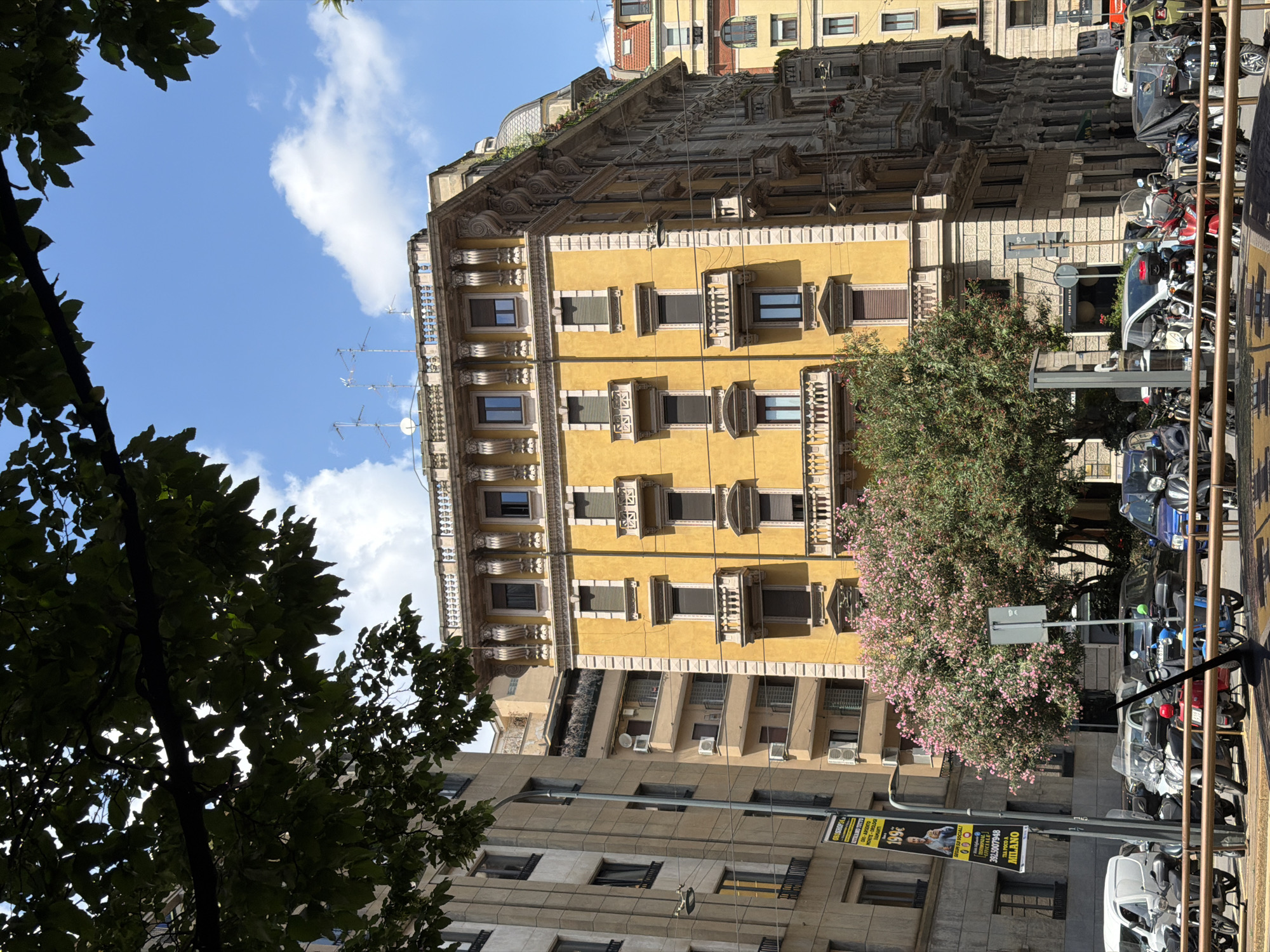 This is a vibrant daytime street scene captured in Milan, Italy, depicting a blend of historic and modern urban architecture under a clear blue sky with scattered white clouds. The main subject is an ornate, multi-story yellow building standing prominently in the upper-middle section of the frame. This building features numerous windows with exterior blinds, decorative balconies, intricate cornices, and architectural embellishments, including satellite dishes on its facade and roof.
Behind the yellow structure, part of a more classical, grey building with a detailed facade and a hint of a rooftop garden is visible. To the left, a more contemporary grey building with simpler lines and visible air conditioning units forms the foreground.
The street level on the right is bustling with parked vehicles, primarily motorcycles and scooters, along with a few cars, characteristic of Italian cityscapes. A metal railing runs along the right edge of the street.
Lush greenery is scattered throughout the scene. A large, dark green leafy tree occupies the lower-left foreground, framing the shot from below. Another smaller tree or bush adorned with delicate pink blossoms adds a touch of color near the yellow building and the street.
Visible text includes an advertisement banner on the left, which reads "199€" and "MILANO", along with smaller Italian text suggesting an exclusive offer for internet and telephony subscribers. A street sign near the pink-flowered tree has "INFO" visible, possibly indicating local information or regulations. The bright sunlight casts distinct shadows, suggesting it's mid-day. No people are visible, lending a quiet quality to this urban snapshot.