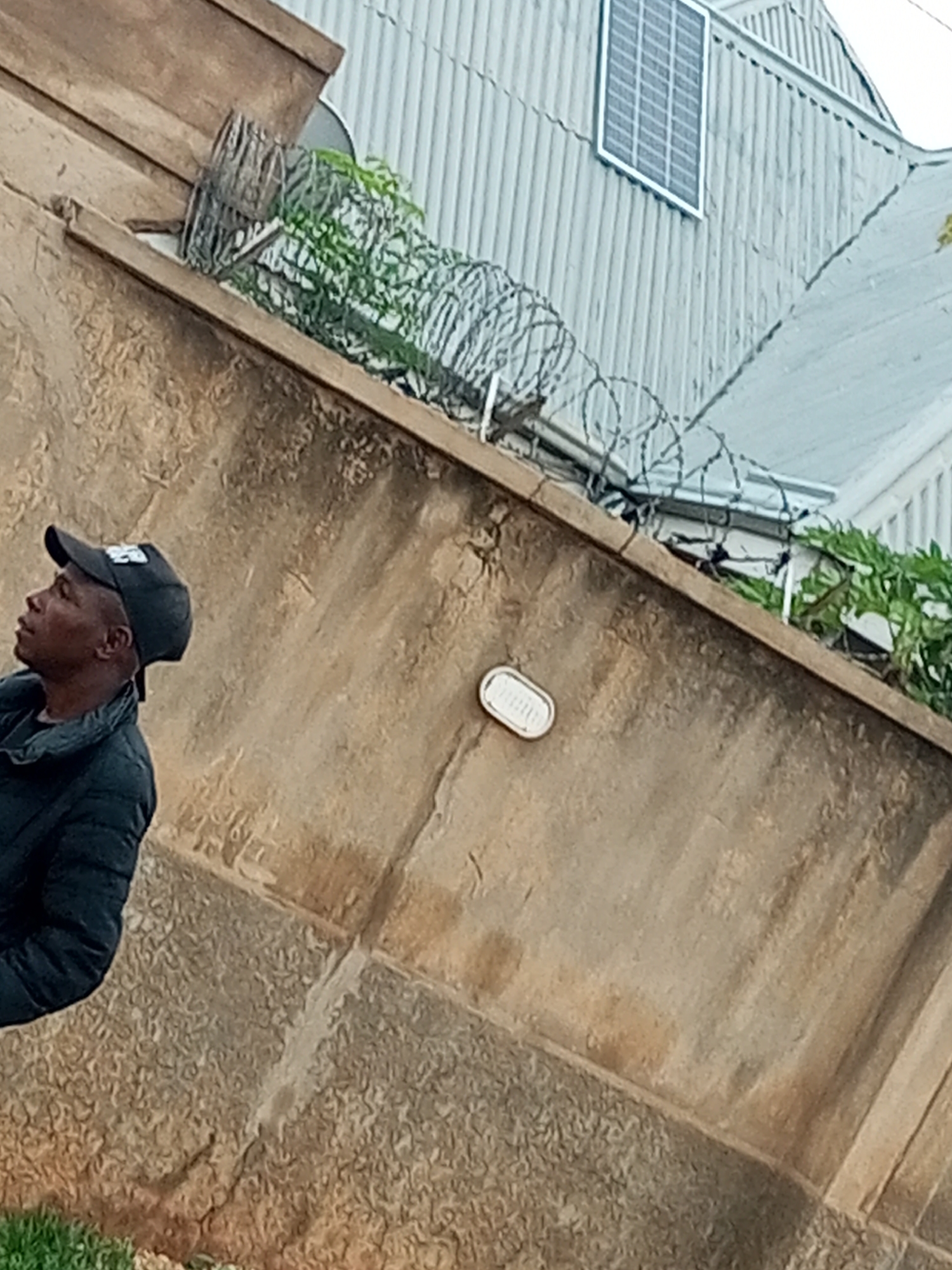 A man wearing a black baseball cap and a dark jacket stands to the left of the frame, facing left and looking upwards. He is positioned against a textured, tan-colored wall. Above the wall, a corrugated metal roof is visible, with a solar panel mounted on it. Barbed wire fencing runs along the top of the wall, interspersed with green foliage. The environment suggests an outdoor setting, possibly a residential or industrial area in Tempure, Nigeria, given the presence of the solar panel and barbed wire, which can indicate security measures. The lighting suggests it is daytime, with the sun illuminating the scene from the upper right.