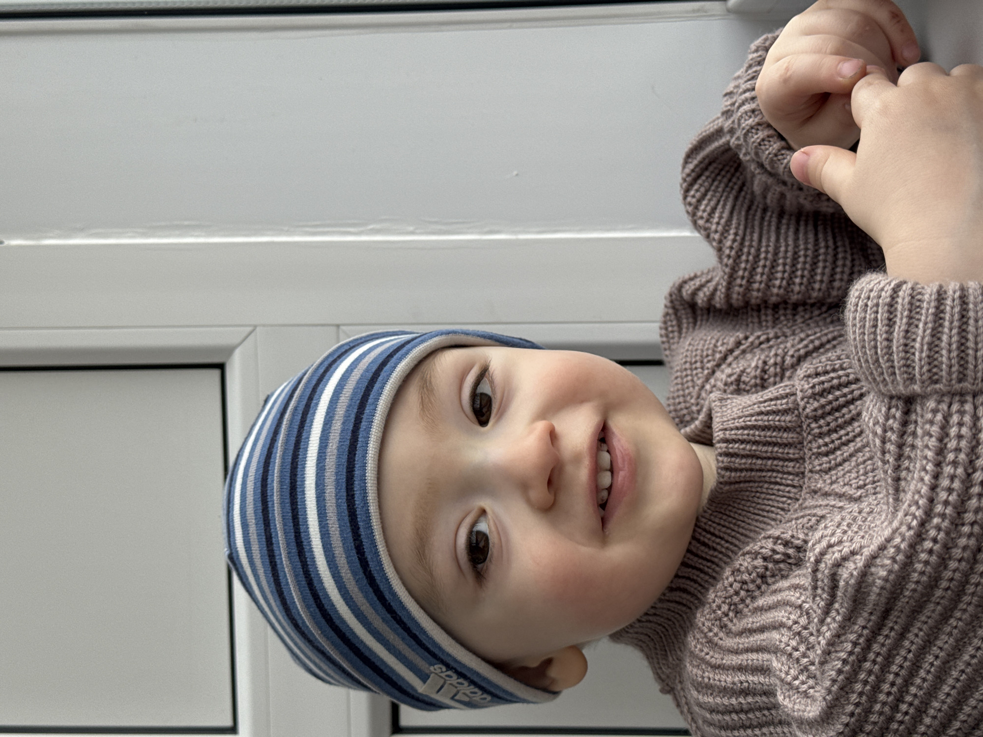 A toddler, wearing a blue and white striped beanie and a brown sweater, smiles at the camera. The child's hands are visible, pressed against a white surface. The location is not identifiable as Izhevsk, Russia, and there are no specific features to determine a precise geographic location.