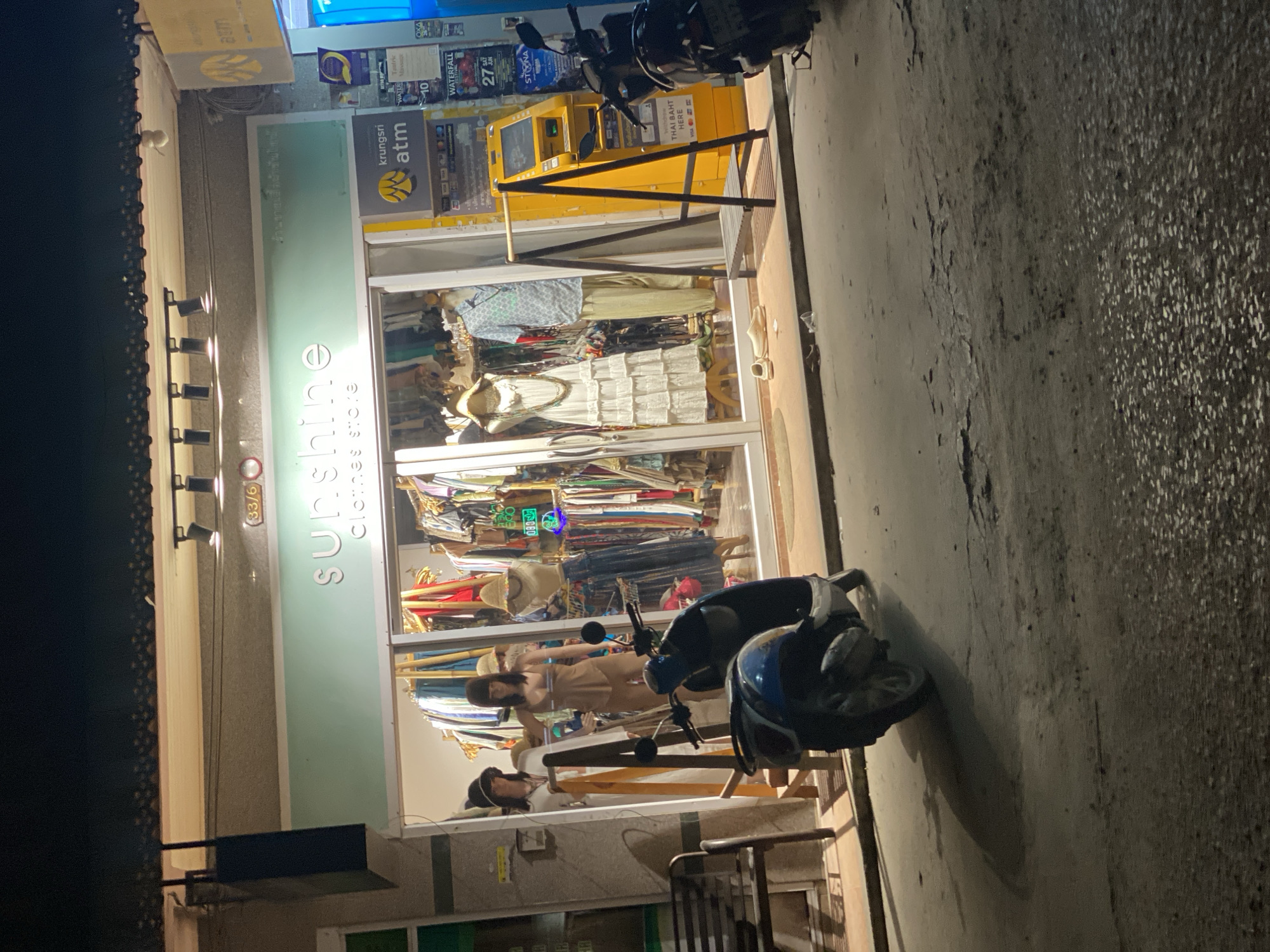 The image shows a nighttime scene outside a clothing store. The store's sign reads "sunshine clothes store" and is illuminated. Inside the store, mannequins are displaying clothes, and racks of clothing are visible. To the right of the store is a yellow ATM machine with signs for "krungsri atm" and "THAI BAHT HERE". A blue and black scooter is parked in front of the store. The ground appears to be a paved street or sidewalk. The lighting suggests it is dark outside, likely nighttime. No specific weather conditions are evident.