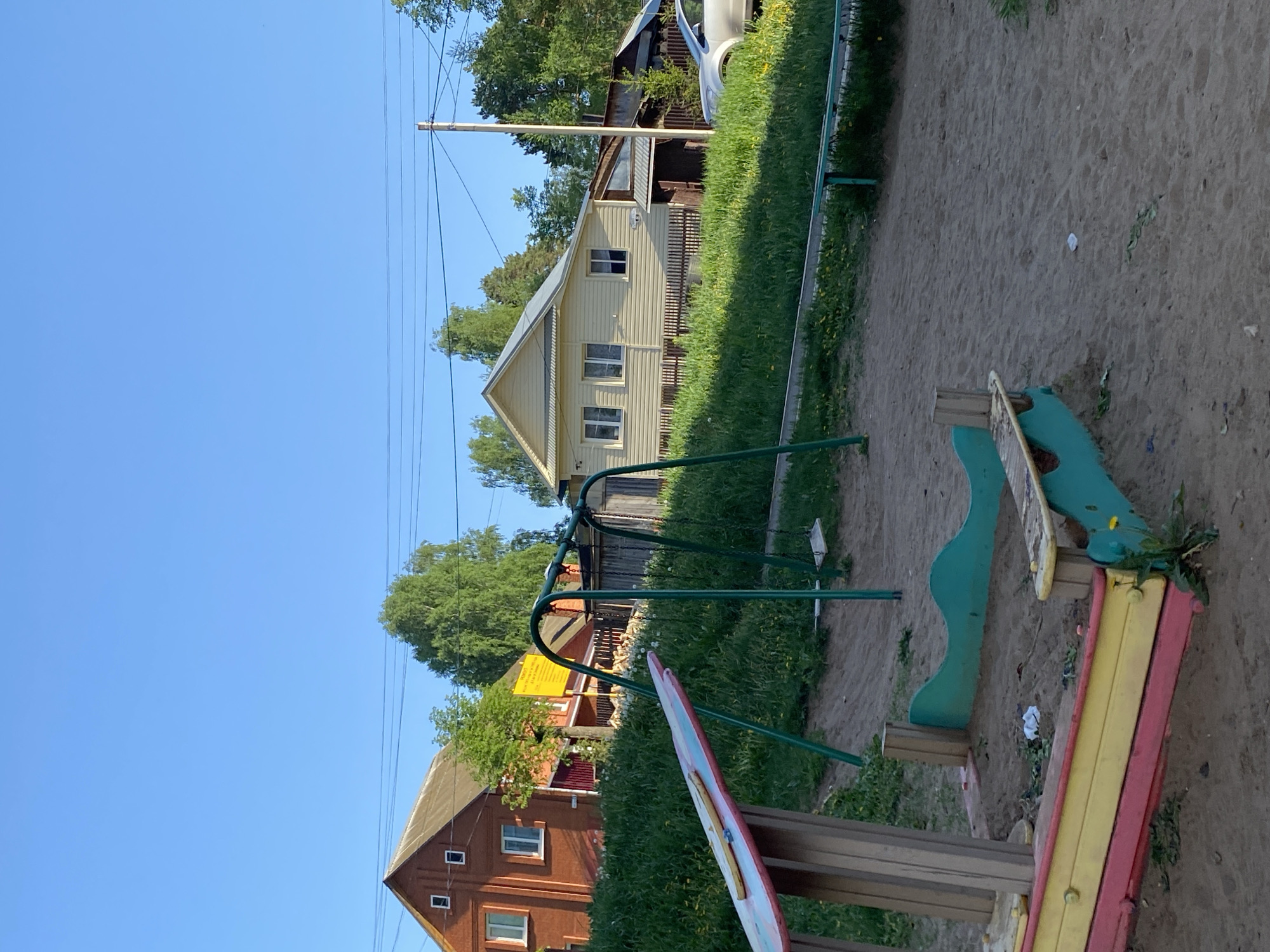 The image shows a playground in Votkinsk, Russia. The playground has a swing set and a rocking horse type of ride. The ground is covered in sand. In the background, there are several houses, including one brick house and one beige house. A power line runs across the sky. The sky is blue.