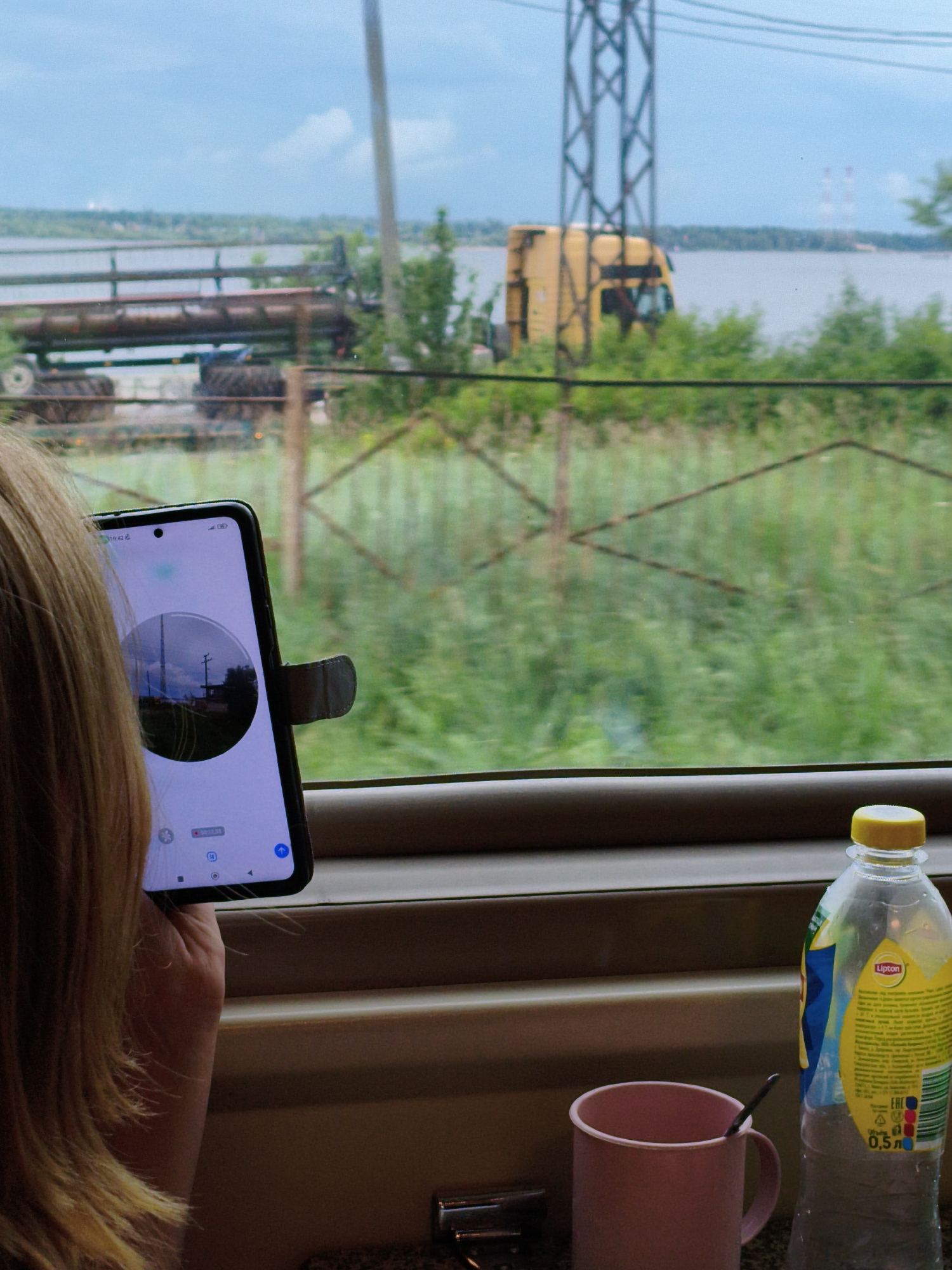 A young person with blond hair is holding a smartphone with a dark case, viewing a circular image on the screen. The phone is positioned in front of a window, through which a landscape is visible. Outside the window, there is a body of water, greenery, a yellow truck, and industrial structures in the distance. A bottle of Lipton iced tea and a pink mug with a spoon are on a ledge below the window. The scene appears to be captured from inside a moving vehicle, possibly a train, judging by the window frame and the blurry background. The overall impression is that of a journey through a semi-industrial, rural or suburban area.