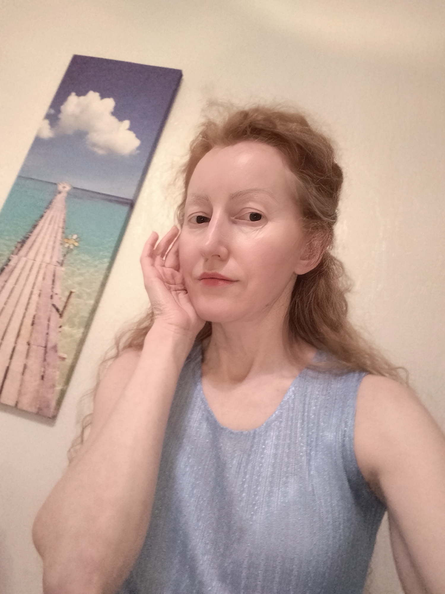 A pale-skinned woman with reddish-blonde hair is posing for a selfie indoors. She is wearing a light blue sleeveless shirt. Her left hand is raised to her face, fingers touching her cheek. Behind her and to the left is a framed picture of a pier extending into a turquoise sea beneath a partly cloudy sky. The wall behind her is light beige. There is no evidence indicating that this image was taken in Krykhivtsi, Ukraine.