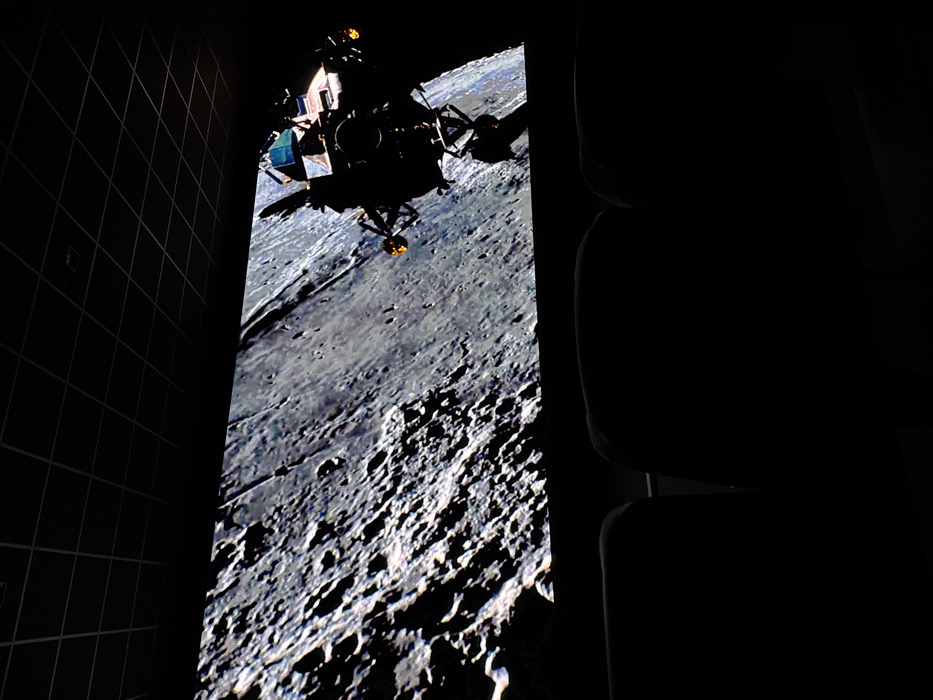 The image captures a forensic view inside an immersive theater or planetarium, specifically the Space Adventure Canela in Canela, Brazil. The dominant feature is a wide, large projection screen displaying a detailed, monochrome depiction of the heavily cratered lunar surface, which occupies the upper two-thirds of the frame. In the upper right quadrant of the screen, the silhouette of a Lunar Module (LM) is visible against the lunar backdrop, its complex structure, landing gear, and antennae clearly discernible. Faint, localized orange-yellow glows are present near the ends of some of the LM's protrusions, possibly indicating thruster activity or reflective surfaces. The room is almost entirely dark, with illumination provided solely by the screen's projection. In the lower third of the image, the dark, featureless silhouettes of two upholstered theater-style seats are visible from a rear perspective, positioned for viewing the screen. The ceiling above is dark with a distinct grid pattern, characteristic of acoustic ceiling tiles in a public venue. No individuals are present or visible within the scene. The overall environment suggests an immersive educational or entertainment experience focused on space exploration.