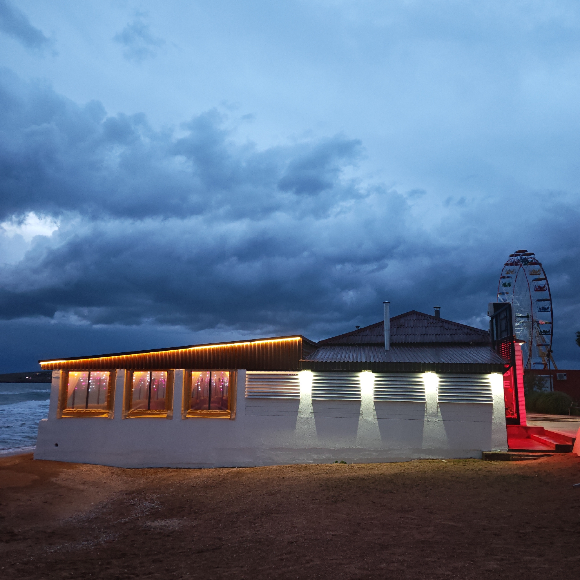 The image depicts a coastal scene at dusk in городское поселение Щелкино, Ukraine. A low, white building stands on the beach, consisting of two sections: one with a flat roof and illuminated string lights above four windows, and another with a gabled roof and exterior lights. The windows of the flat-roofed section appear to have curtains or decorations inside. To the right of the building, a lit Ferris wheel is partially visible. The sky is filled with dark, heavy clouds, indicating an approaching storm. The ground is dark and appears to be sand. The ocean is visible in the background to the left of the building. There are no people visible in the image.