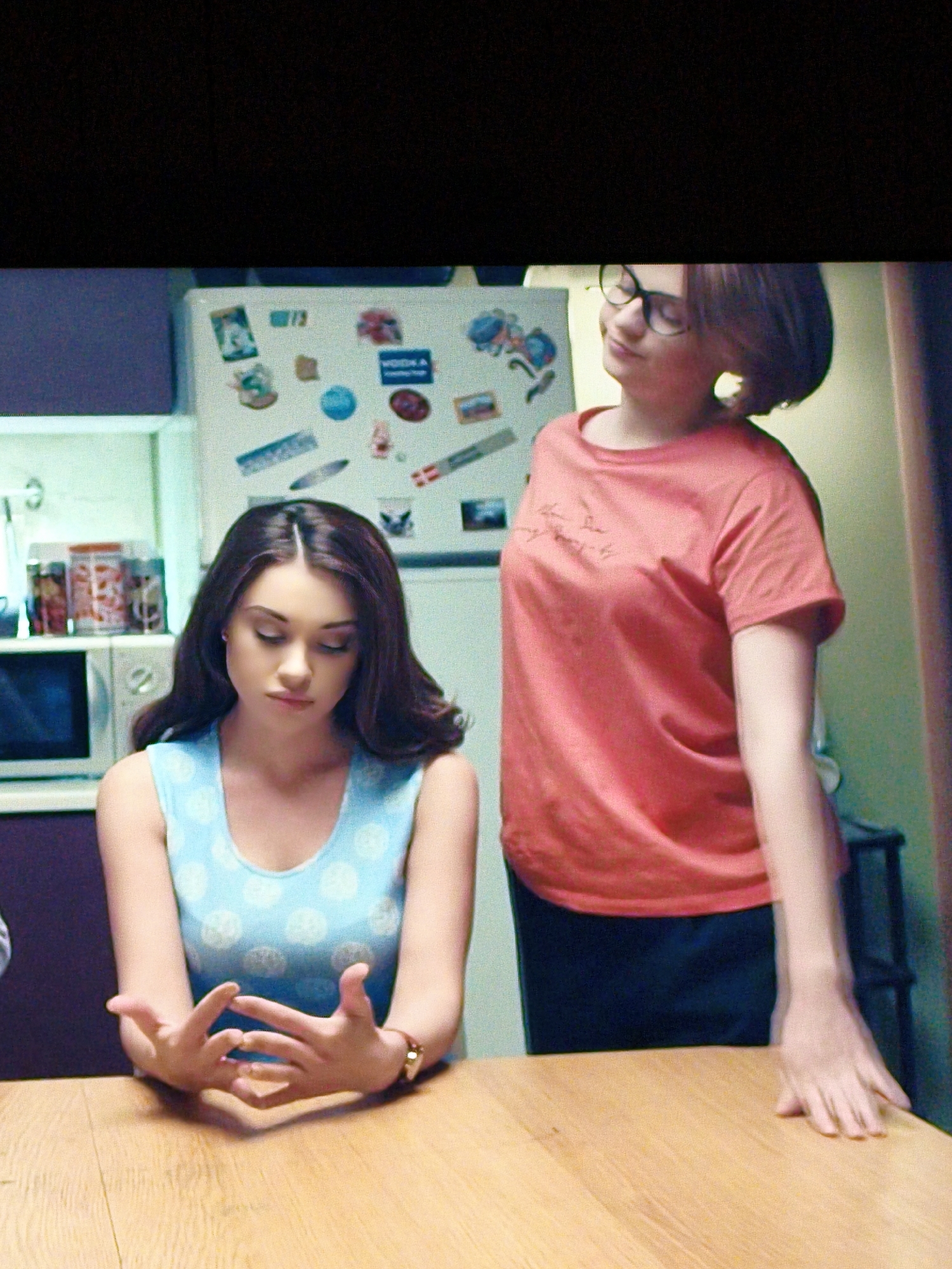 The image depicts two young women in a kitchen. The woman on the left, with dark, wavy hair, is wearing a light blue dress with a white polka dot pattern. She is seated at a wooden table, with her hands clasped in front of her and her head bowed. A watch is visible on her left wrist. The woman on the right, with short brown hair and glasses, is wearing a bright coral-colored t-shirt and dark pants. She is standing and appears to be speaking or looking at something to her left. Behind the women, a white refrigerator is covered in various magnets and papers. A microwave and shelves with jars are visible to the left of the refrigerator. The kitchen appears to be in a residential setting. There is no indication of the specific location within Vladivostok, Russia.