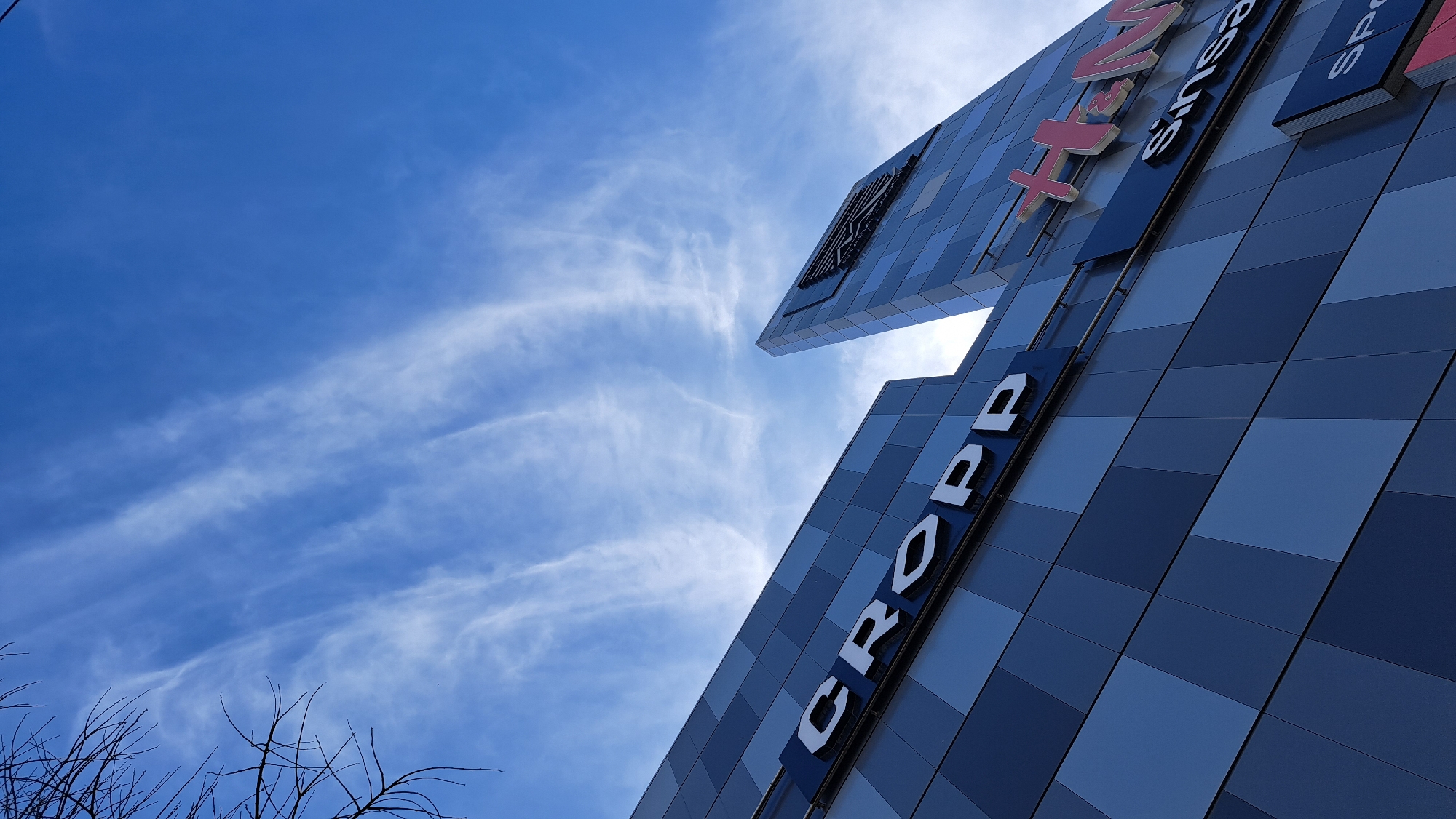 A low-angle shot captures the modern facade of a building against a vibrant blue sky with wispy white clouds. The building's exterior is constructed from an array of square and rectangular panels in varying shades of blue and gray, creating a mosaic-like pattern. Large, three-dimensional white letters spell "CROPP" prominently on the lower section of the building, positioned diagonally. Further up and to the right, partial signage for other retail brands is visible, including red letters spelling "H&M" and white letters for "Sinsay," along with an additional "SP" sign. These visible texts suggest the building is part of a shopping center or retail complex.
The sun is bright, creating a slight glare near the top right, indicating it is daytime, likely a sunny morning or afternoon. Bare tree branches are visible in the bottom left corner, hinting at a season without foliage, such as late autumn or winter. There are no people visible, and no specific activity is taking place. The overall impression is one of modern architecture and commercial activity under a clear, open sky.