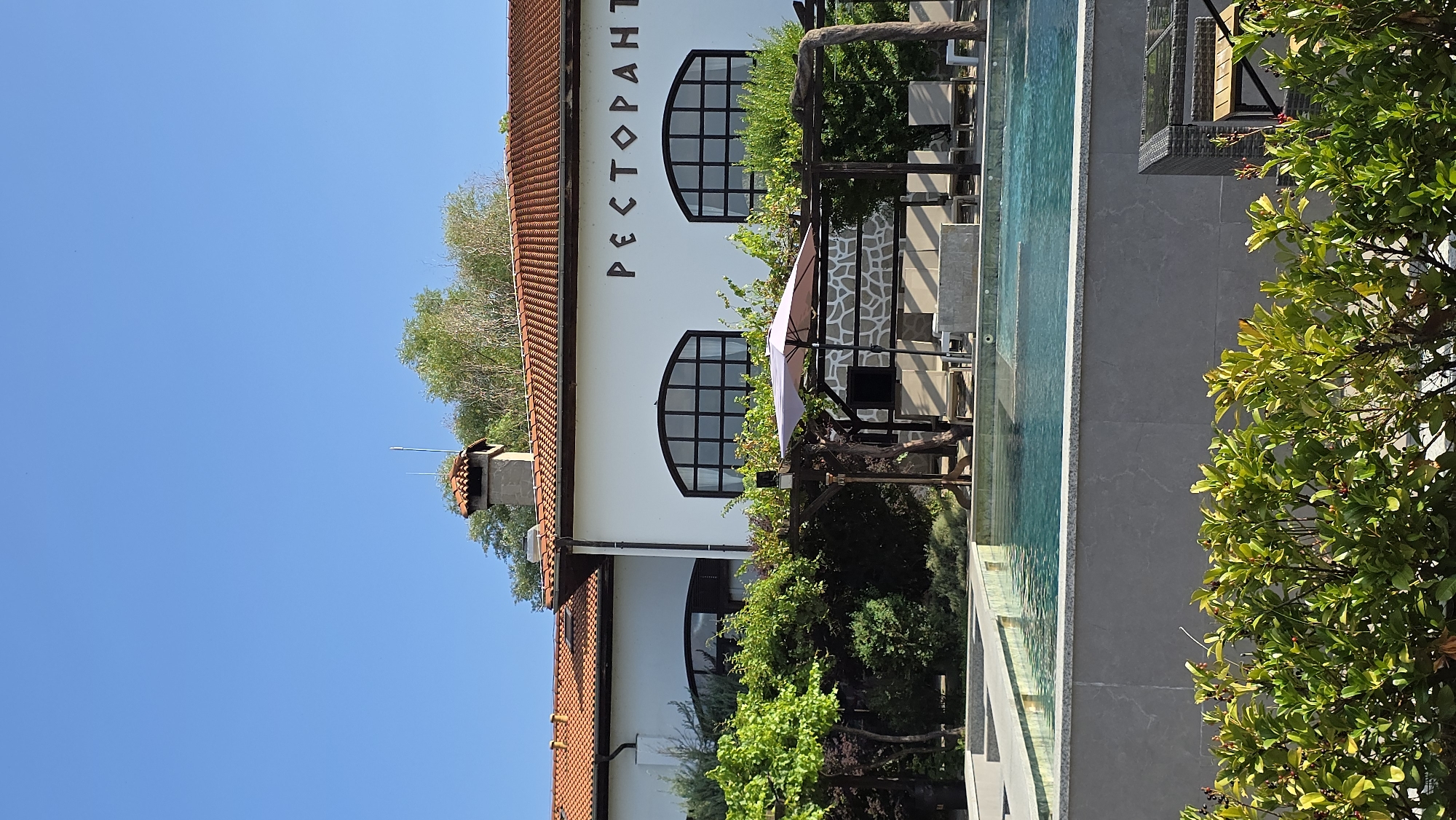 In Krasnovo, Bulgaria, a picturesque scene unfolds at a resort. A swimming pool, reflecting the clear blue sky, takes center stage, bordered by a textured gray wall. Beyond the pool, lounge chairs and an umbrella suggest a place for relaxation. A pergola with intertwined vines frames the view, leading to a white building adorned with arched windows and the word "РЕСТОРАНТ" (Restaurant) in large, dark lettering. The building features a traditional red-tiled roof and a chimney. Lush green foliage, including vines and bushes, surrounds the area, creating a vibrant and inviting atmosphere.