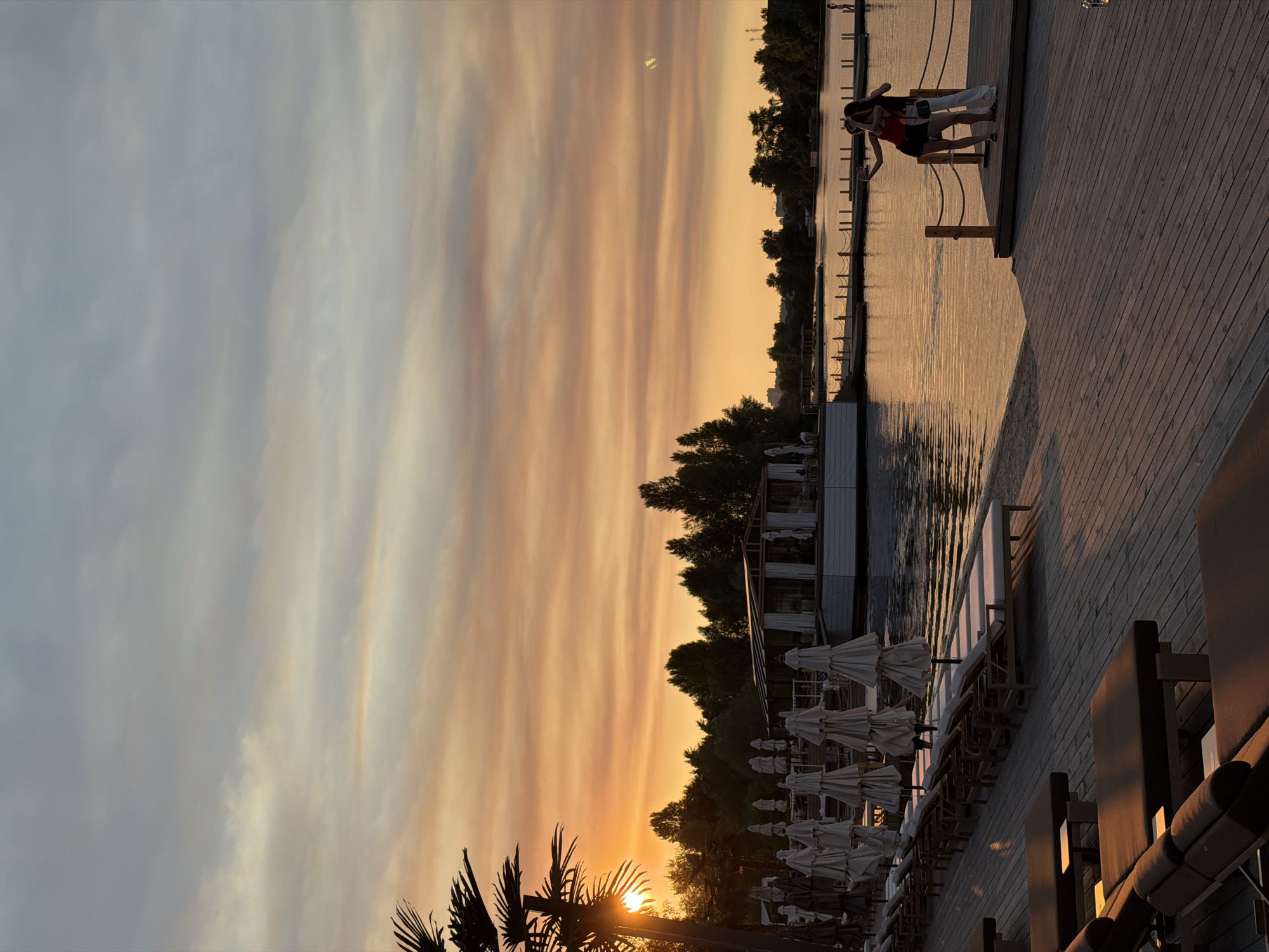 The image depicts a serene sunset over a body of water, likely a river or lake, in Kyiv, Ukraine. Two individuals, a person wearing a red top and black shorts, and another person in a white top and pants, are standing on a wooden pier, engaged in conversation or observation. The sky is painted with streaks of orange and yellow from the setting sun, with hints of gray and blue clouds.
The pier is lined with closed beach umbrellas and empty lounge chairs, suggesting the end of a day at a beach club or resort. Palm trees frame the lower left corner of the image, adding a tropical touch to the scene, which is unusual for Kyiv but could indicate a specific establishment with such landscaping. The water reflects the warm hues of the sunset, creating a tranquil atmosphere.
The lighting indicates it is late afternoon or early evening, during the golden hour, known for its soft, warm light. The overall mood is peaceful and contemplative, with the natural beauty of the sunset dominating the scene. There is no visible text in the image.
