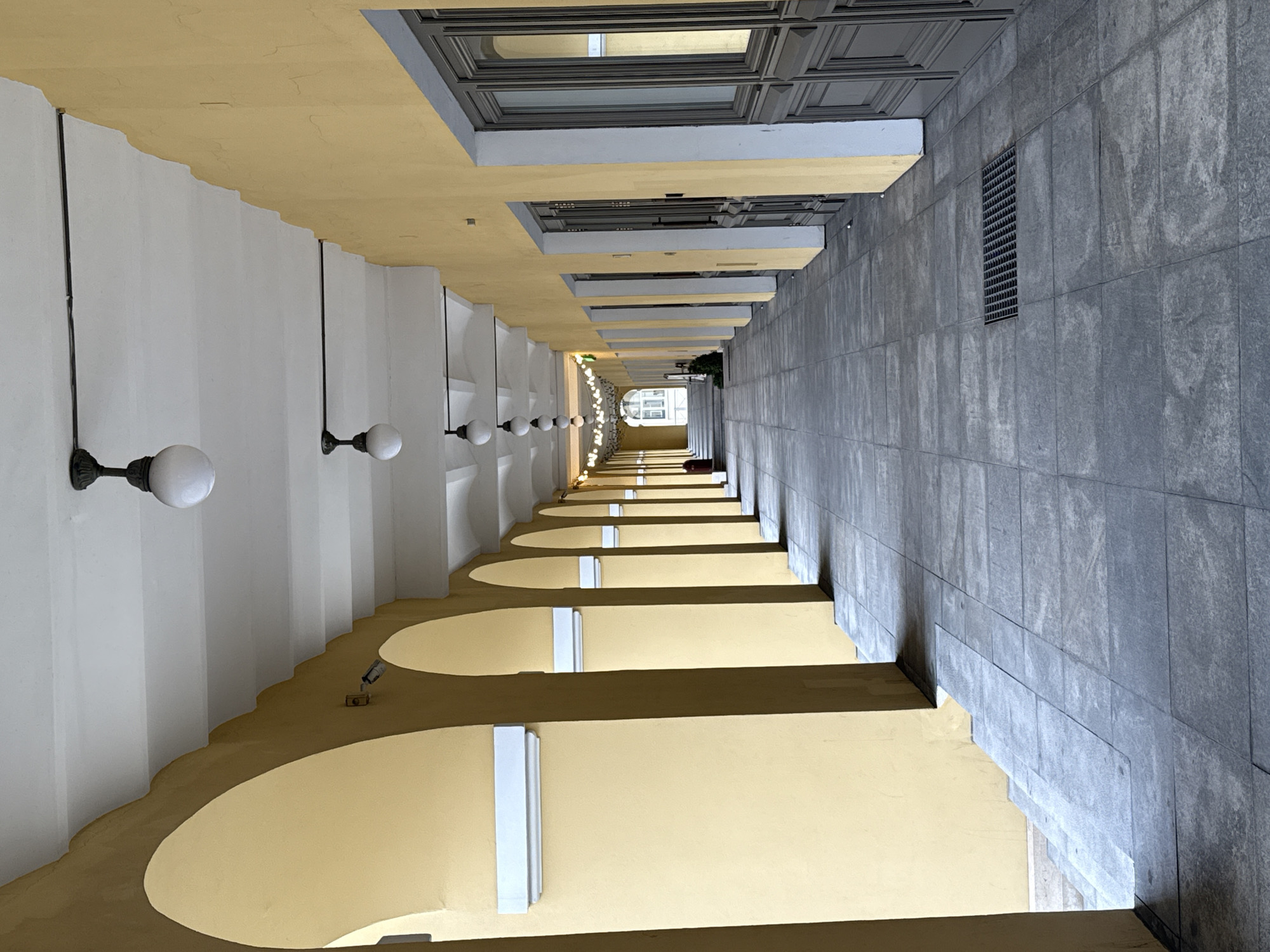This image captures a long, architectural passageway or covered courtyard in Saint Petersburg, Russia, viewed from one end looking down its length. The perspective is strong, emphasizing the depth and repetition of architectural elements.
On the left side, a series of white walls supports light yellow arches, forming a graceful arcade or portico. Attached to the white walls are evenly spaced, antique-style spherical wall lamps with dark metal fixtures, providing illumination. Beneath these arches, the floor is a darker yellow/brown, punctuated by lighter, illuminated circular or oval sections, possibly integrated lighting or skylights.
The right side of the passageway features a grey tiled wall with a prominent rectangular metal grate. Above this wall, a series of recessed window-like openings or decorative panels with grey frames reflect light, suggesting an upper story of a building. The ground on this side is paved with rectangular grey tiles.
The passageway narrows towards the distant end, where string lights are visible overhead, illuminating a brighter area that appears to be an exit or an open courtyard. Some greenery, possibly a small tree or bush, is also visible at the far end.
The lighting appears bright and uniform, suggesting either an overcast day or an interior space with consistent artificial light, although shadows cast by the arches indicate a primary light source from above. There are no people visible, and no discernible activity is taking place. The scene conveys a sense of order, elegance, and quietude. No text is visible in the image.