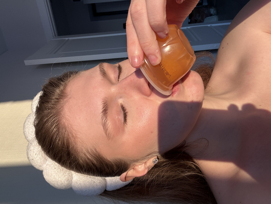 A female is depicted in a close-up, chest-up shot within an indoor setting in Kazan, Russia. She is shown in profile, facing right, with her eyes closed and lips pursed around the rim of a small, clear plastic cup. The cup contains an amber-colored liquid or gel and is held by her right hand. Visible text on the cup reads "5.30-04.21" and "3700-01.31". She wears a white, puffy headband and a round, clear stud earring in her visible left earlobe. Her shoulders and upper chest are bare. Strong, warm light illuminates her face from the upper left, casting a prominent shadow across her right cheek, jawline, and neck. The background is out of focus, revealing a light-colored wall and a white door frame with a partially open white door and a dark interior to the upper right.