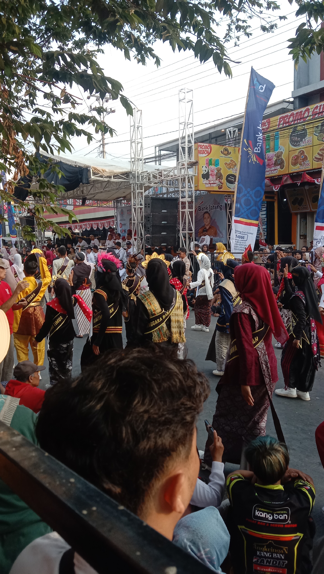 A crowd watches a parade in Jatisawit, Indonesia. In the foreground, a person with dark curly hair is looking away from the camera. Behind them, another person with blue-tinted hair is seated, wearing a black shirt with yellow lettering. The parade participants, dressed in colorful traditional attire with sashes, walk in the street. In the background, a stage is set up with speakers and scaffolding, and banners for "PROMO HEBOH" and "Bank Jateng" are visible on buildings. Trees with green leaves hang over the scene, casting shadows.
