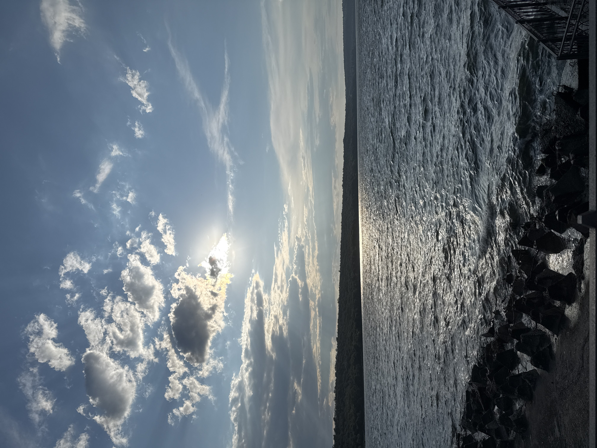 The image depicts a serene natural scene with a body of water stretching towards a distant, tree-lined shore under a partly cloudy sky. The sun is visible, partially obscured by clouds, casting a shimmering reflection on the water's surface. In the foreground, a dark, rocky embankment lines the water's edge, with a metal railing visible to the right, suggesting a public access point or a viewing area. The overall impression is one of tranquility and natural beauty, likely captured during the late afternoon or early evening. The location is specified as the city of Korzhevo, Moldova. There are no people or animals present in the image, and no specific actions or interactions are depicted. The environment is characterized by water, landmass with dense vegetation, and a dynamic sky.