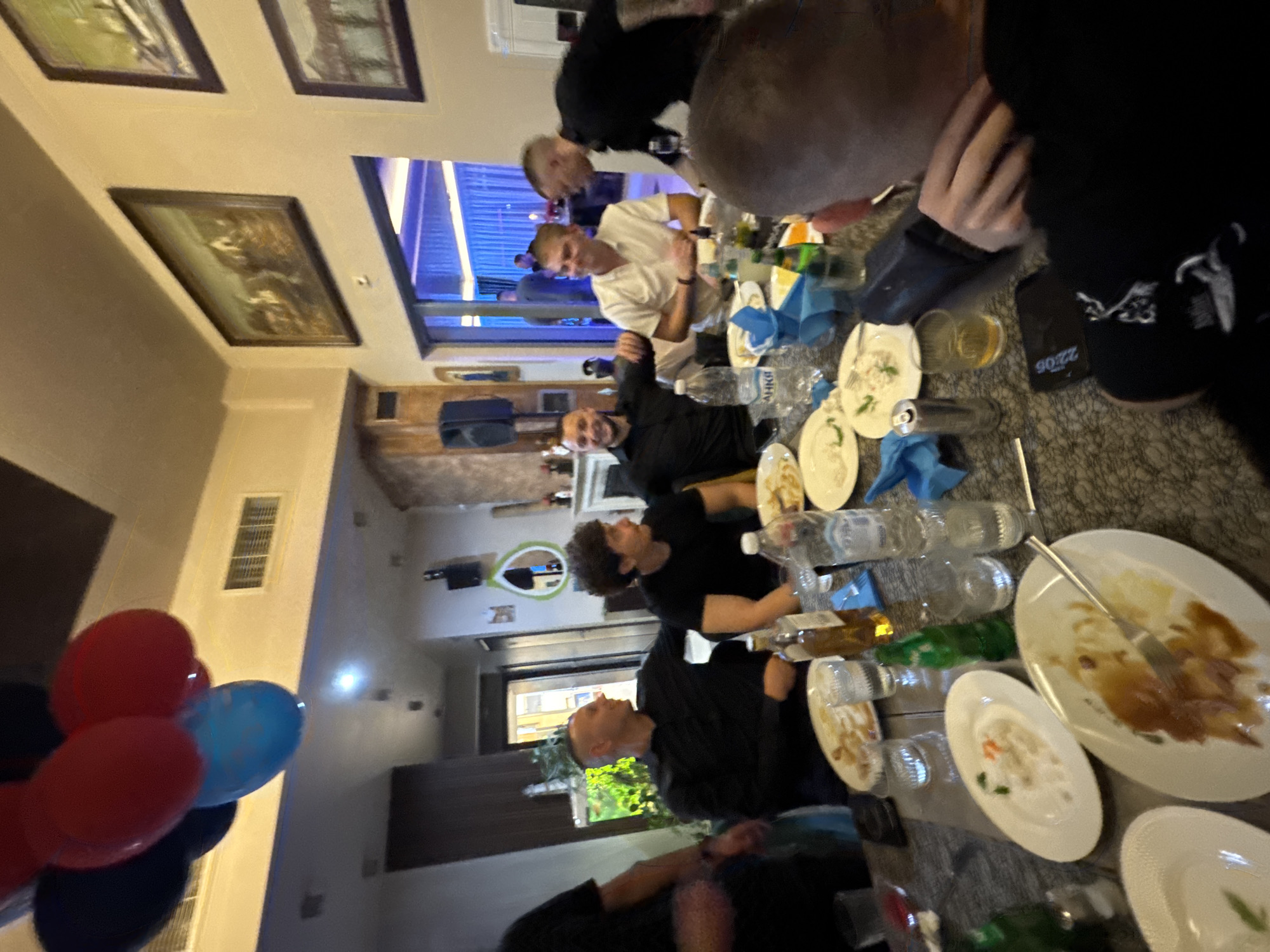 The scene is inside a restaurant or dining area. Several individuals are seated around a table laden with plates, bottles, and glasses. The plates appear to contain remnants of food. Some of the individuals are dressed in black attire. A cluster of red and blue balloons is visible. The location is possibly in Plovdiv, Bulgaria. Three picture frames hang on the wall and an open window looks outside.