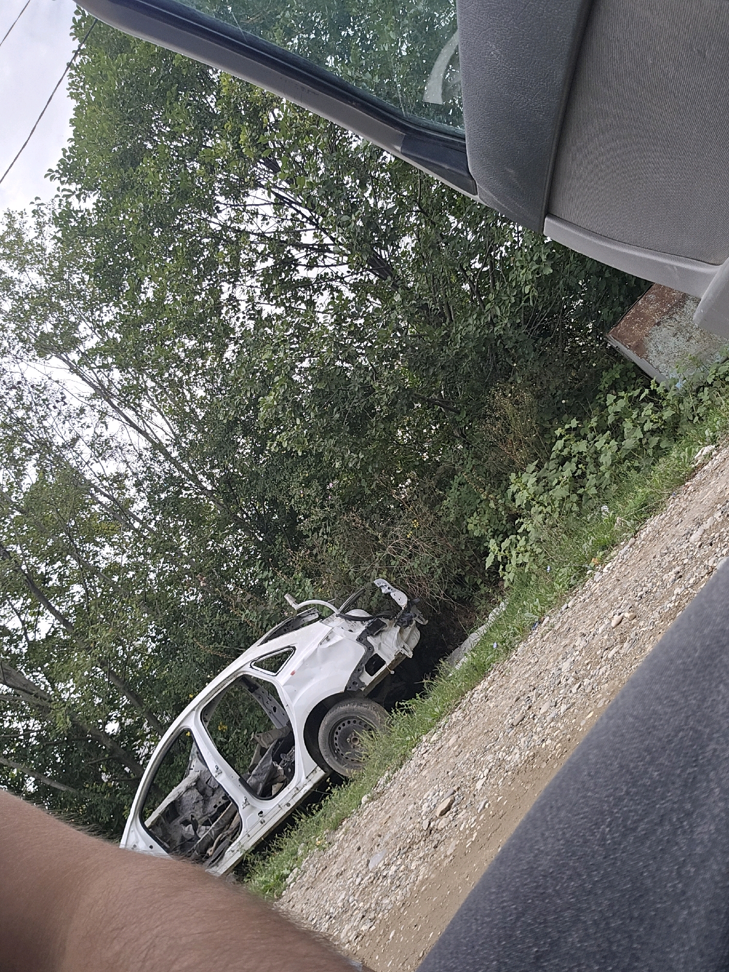 The image shows a white, heavily damaged car body, likely stripped of its internal components, resting on its side next to a gravelly embankment and lush green foliage. The car is missing its doors, hood, and trunk, revealing its skeletal interior. One of the wheels is still attached. The setting appears to be outdoors, with a dense canopy of green trees in the background. The angle of the shot suggests it was taken from inside another vehicle, with a portion of a gray seat and a dark grey fabric-like material in the foreground. The overall scene evokes a sense of abandonment or a junkyard. The location is identified as Avtury, Russia.