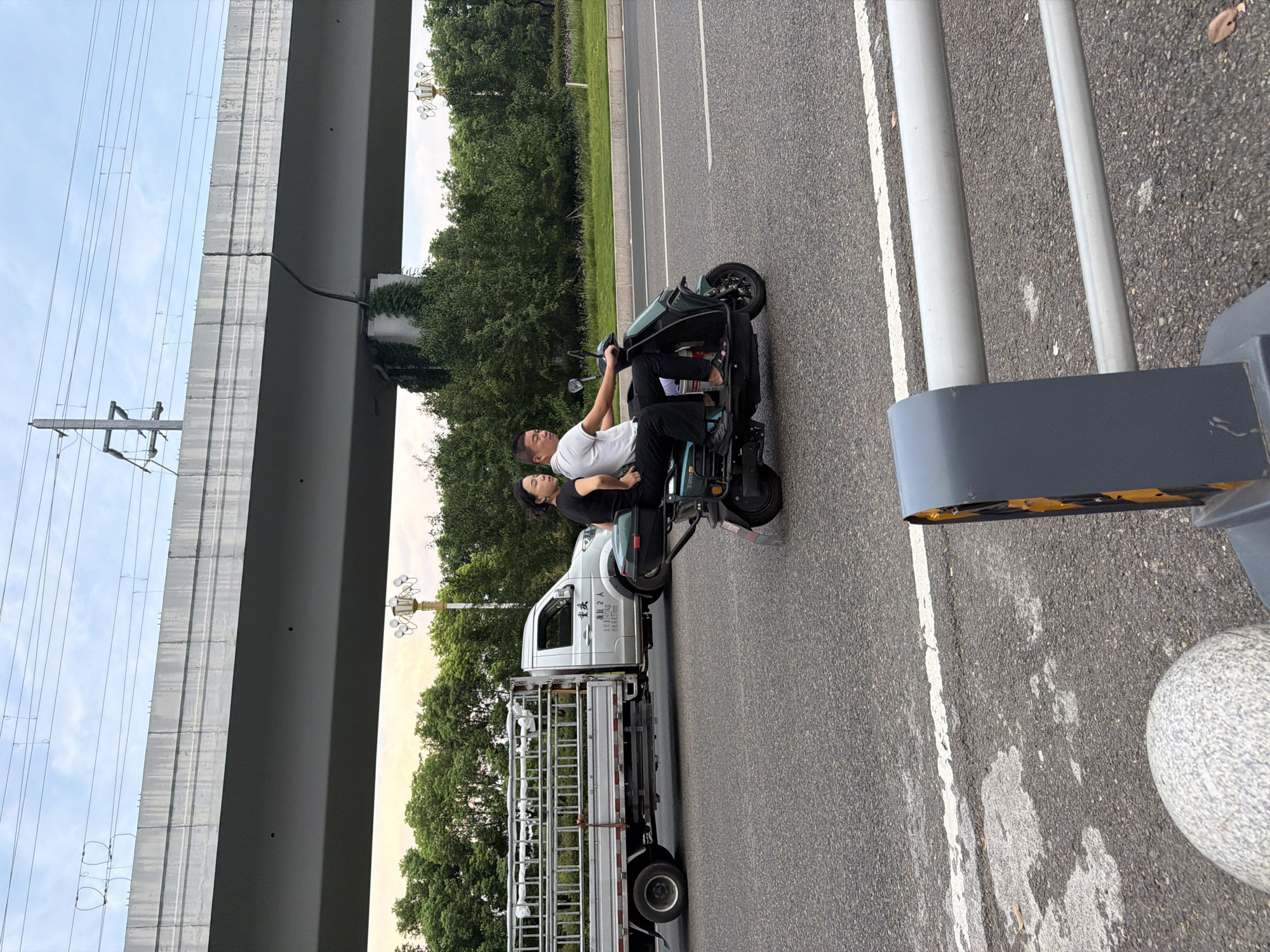 In Jiangjin, China, a man and a woman are riding a scooter on an asphalt road. The man is driving, and the woman is sitting behind him. Both are wearing dark clothing. The scooter is green and has a storage box on the back. To the left of the scooter is a silver flatbed truck with a metal cage structure on its bed. The truck has Chinese characters on its door. Behind the vehicles, lush green trees line the roadside, with a large elevated concrete structure, likely an overpass or bridge, spanning across the background. Power lines are visible above the elevated structure, and a street light with multiple globes stands to the right. The road has white lane markings. The foreground shows a gray concrete bollard and a section of a metal barrier.